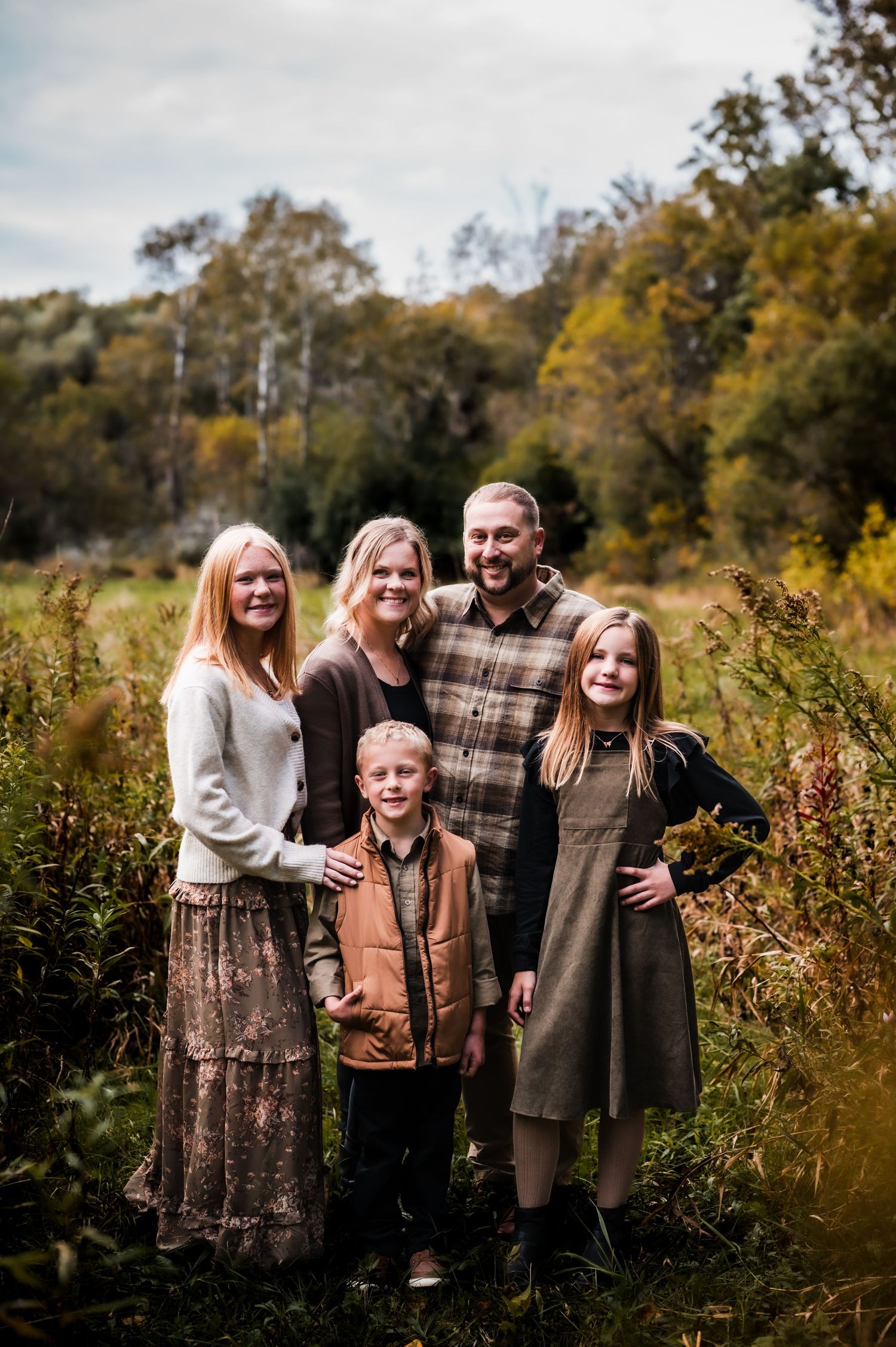 Family of five posing outdoors in autumn field with colorful fall foliage in background.