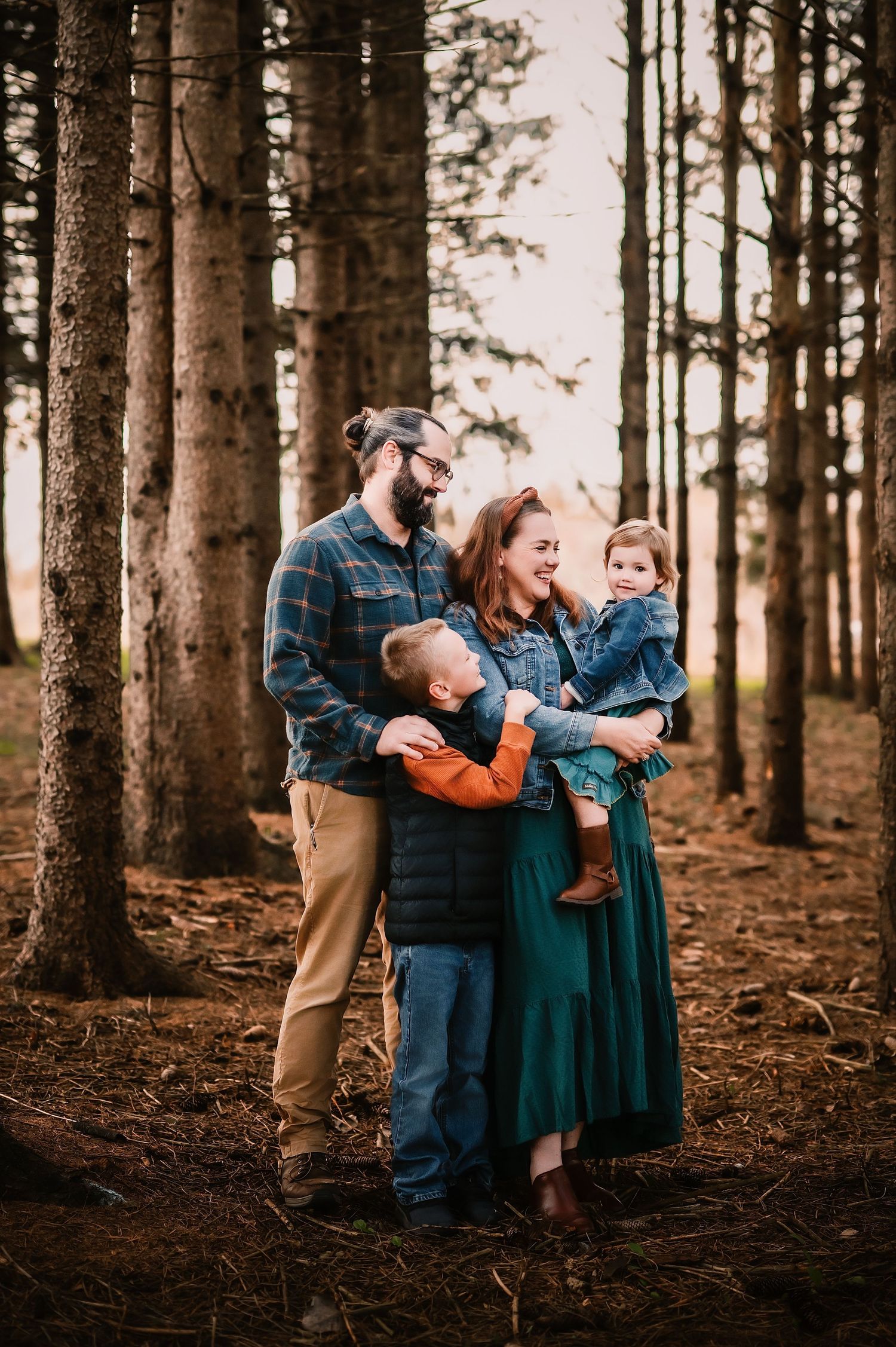Family of four poses in a pine forest, parents smiling as young children embrace them in a warm autumn photo session.