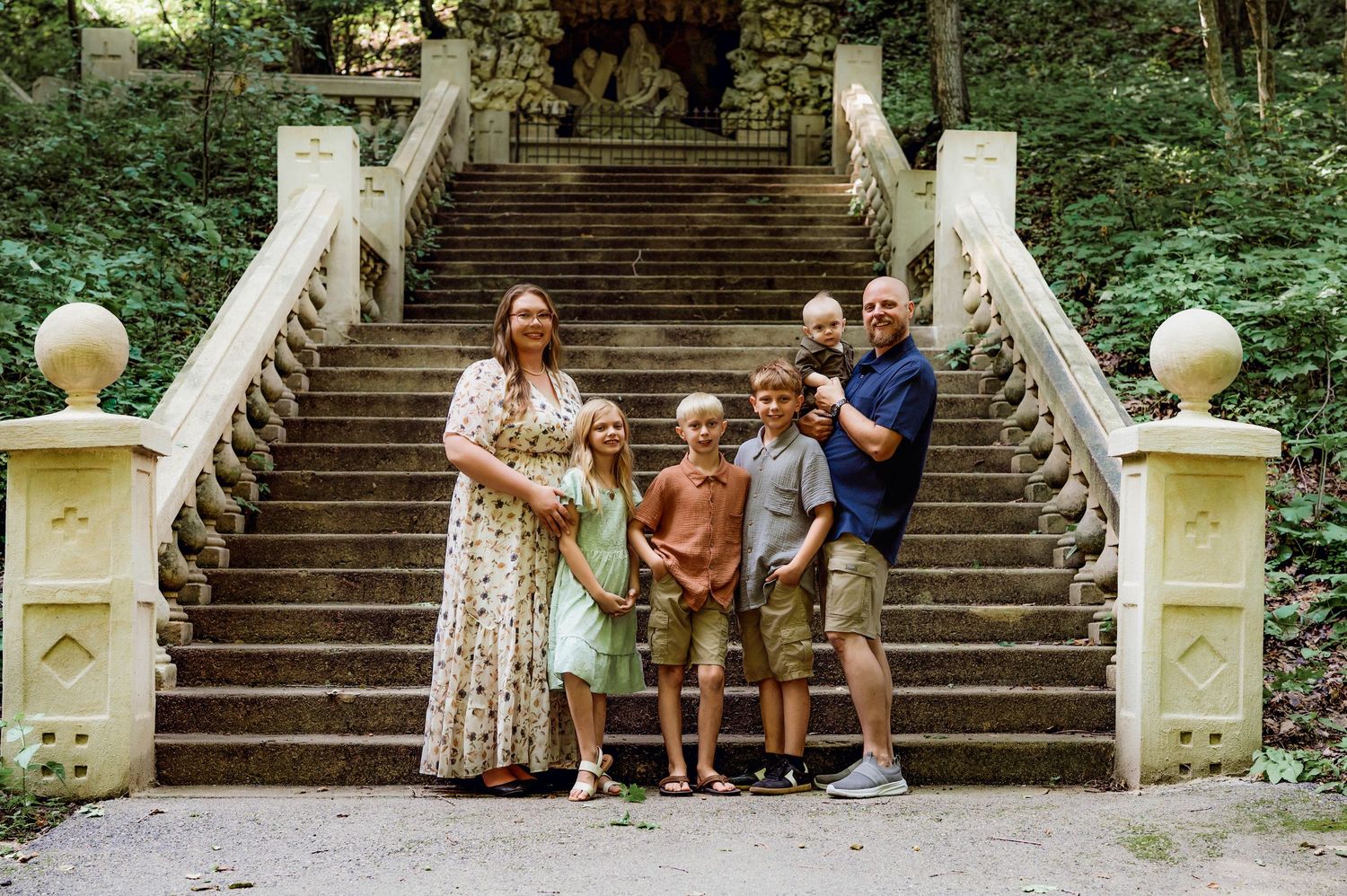 Family of six posing on ornate stone staircase surrounded by lush green trees in a scenic outdoor setting.