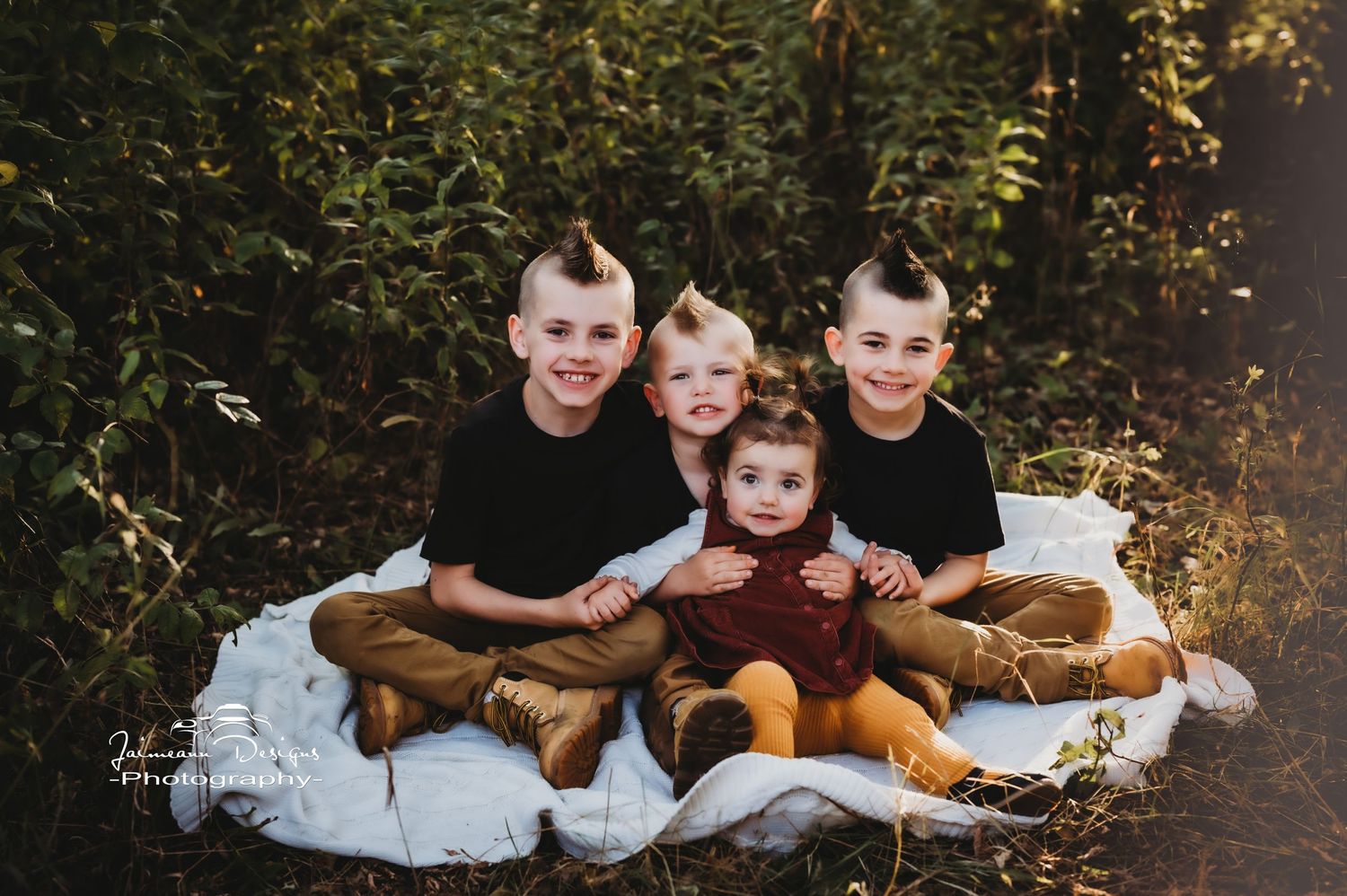 Children sitting together on a white blanket outdoors with autumn foliage in the background.
