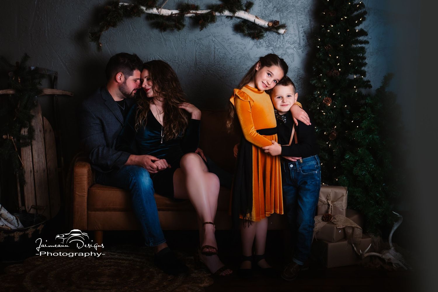 Family sits together on leather couch in moody holiday setting with pine garland and lights.