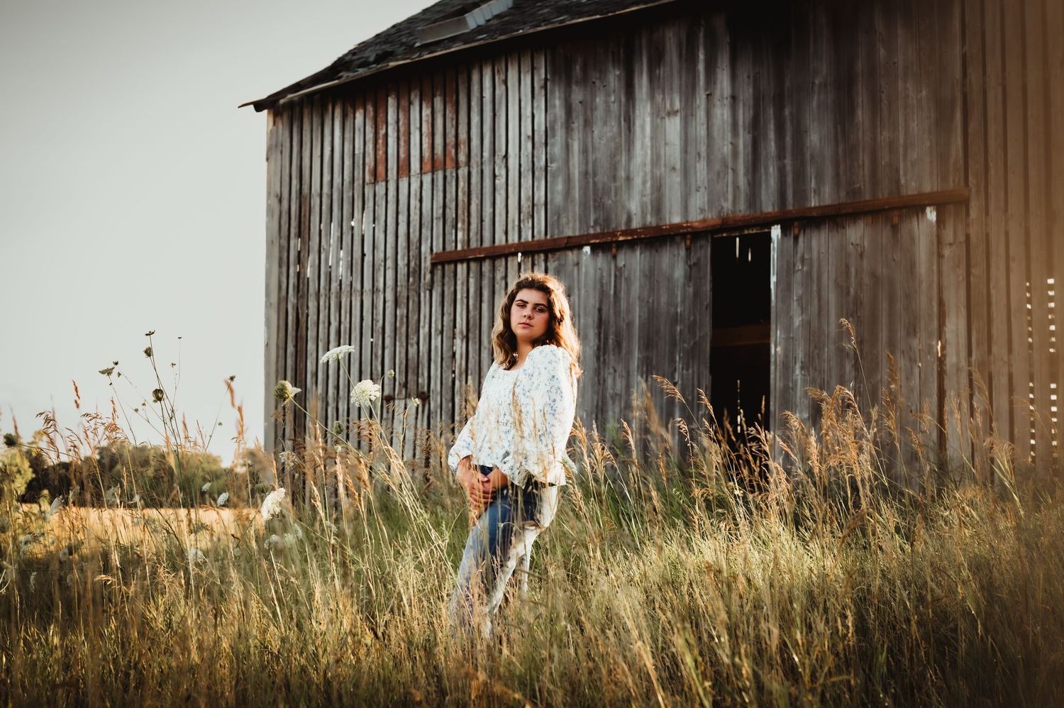 Rustic outdoor portrait with person standing in tall grass near an old wooden barn.