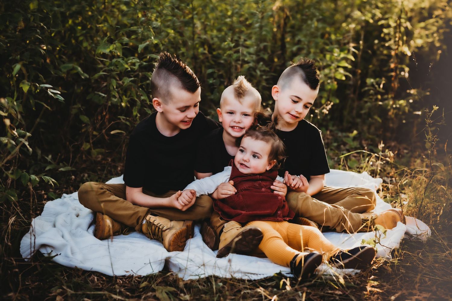 Four youths sit together on a white blanket outdoors wearing casual fall clothing in warm golden light.