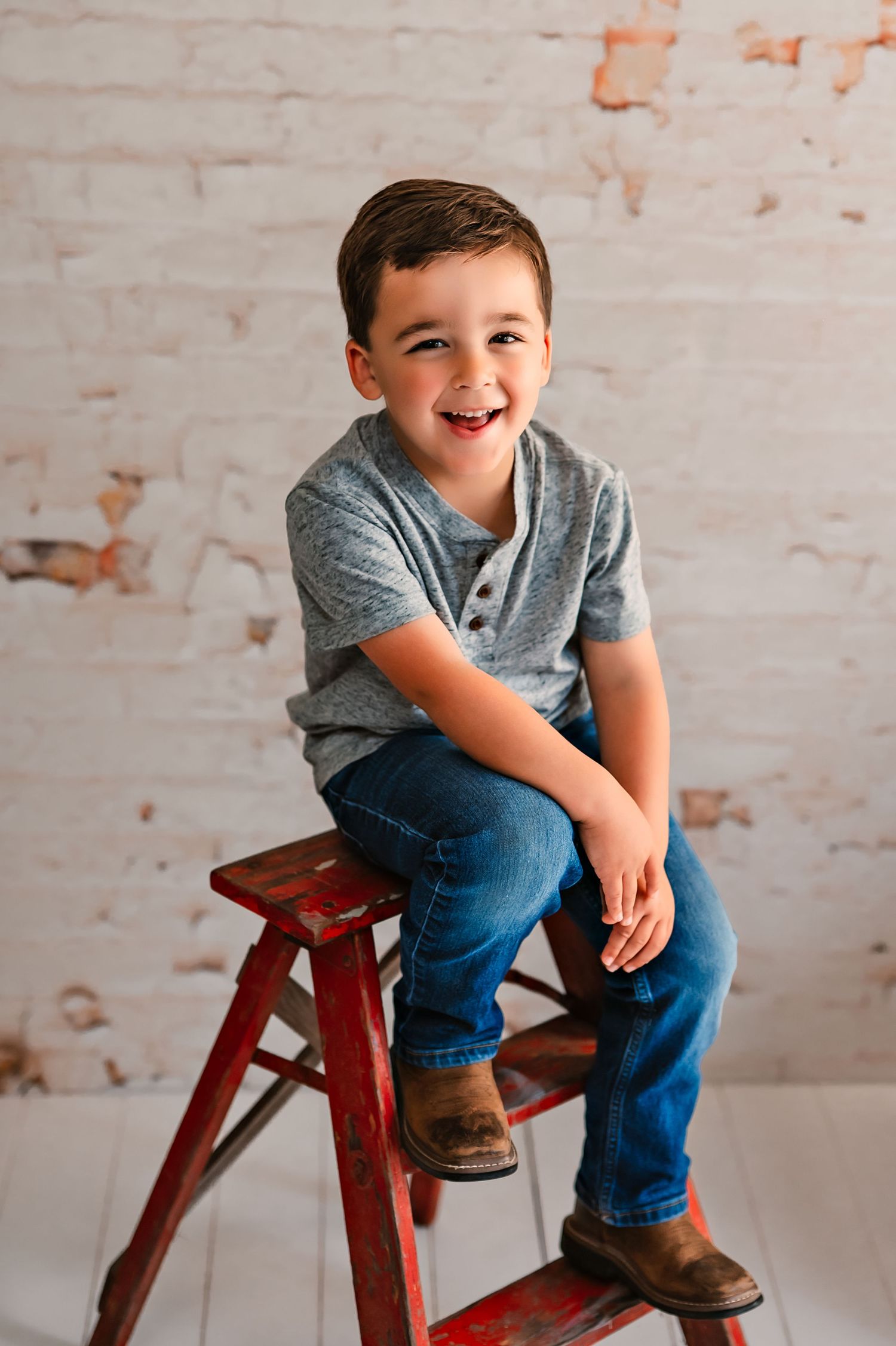 Child wearing blue jeans and gray shirt sits on wooden stool against white brick wall, In Kewaskum, WI Studio