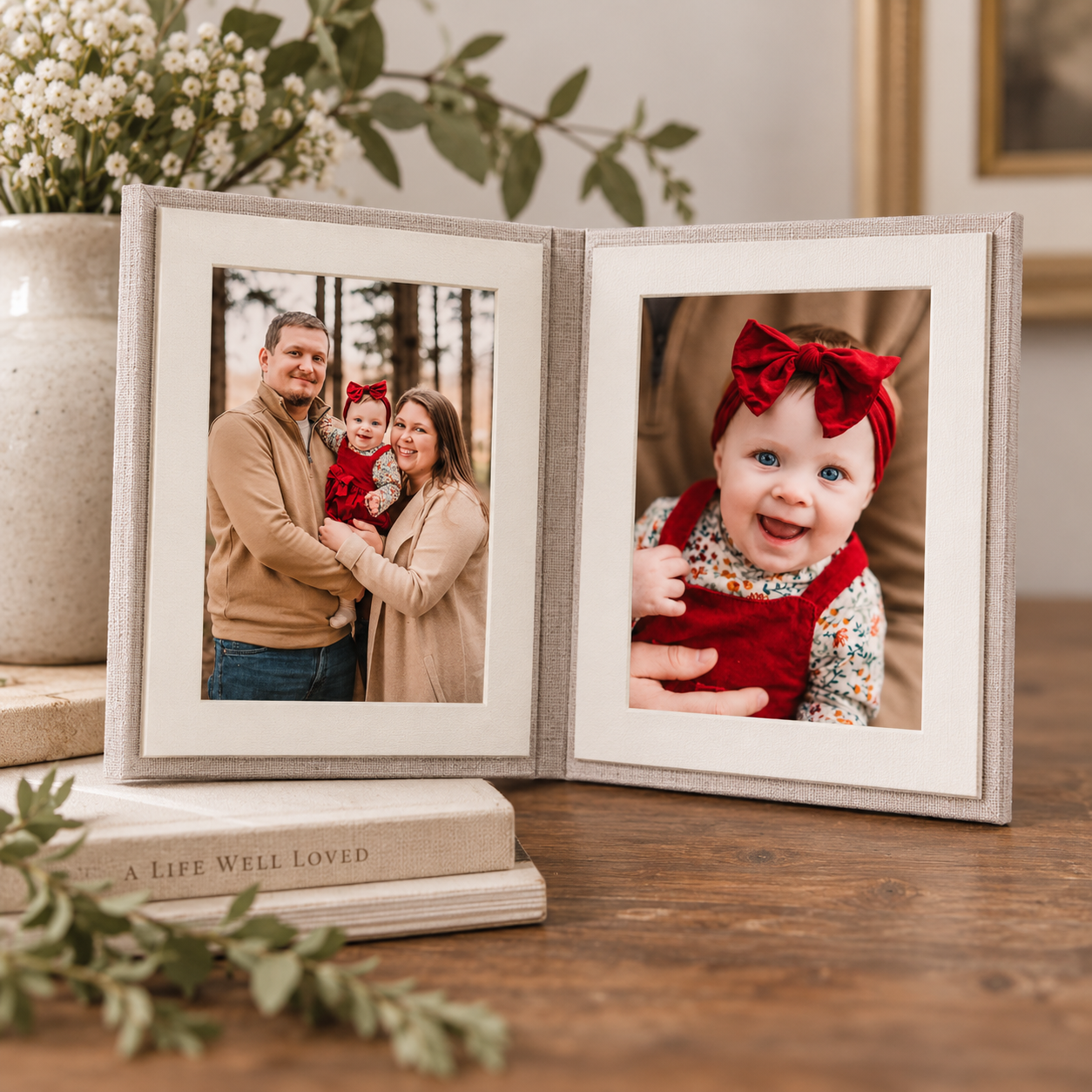 A hinged double picture frame displaying a family portrait and a baby girl in a red bow, sitting on stacked books on a wooden table.