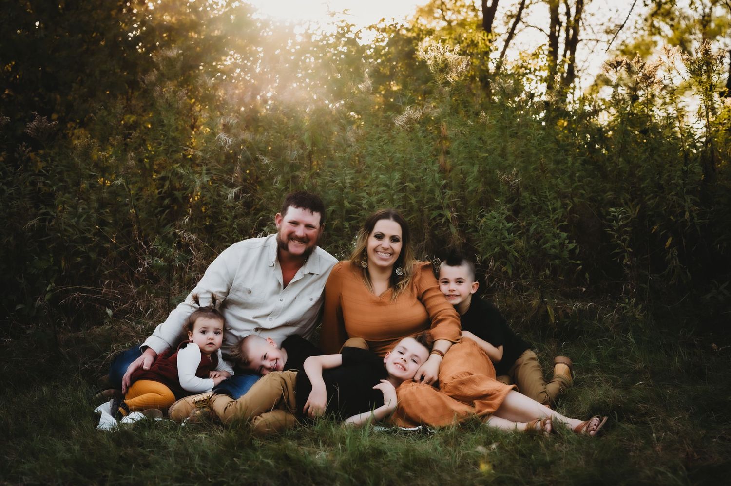 A family sits together on grass during golden hour sunset in autumn, wearing coordinating warm-toned outfits.