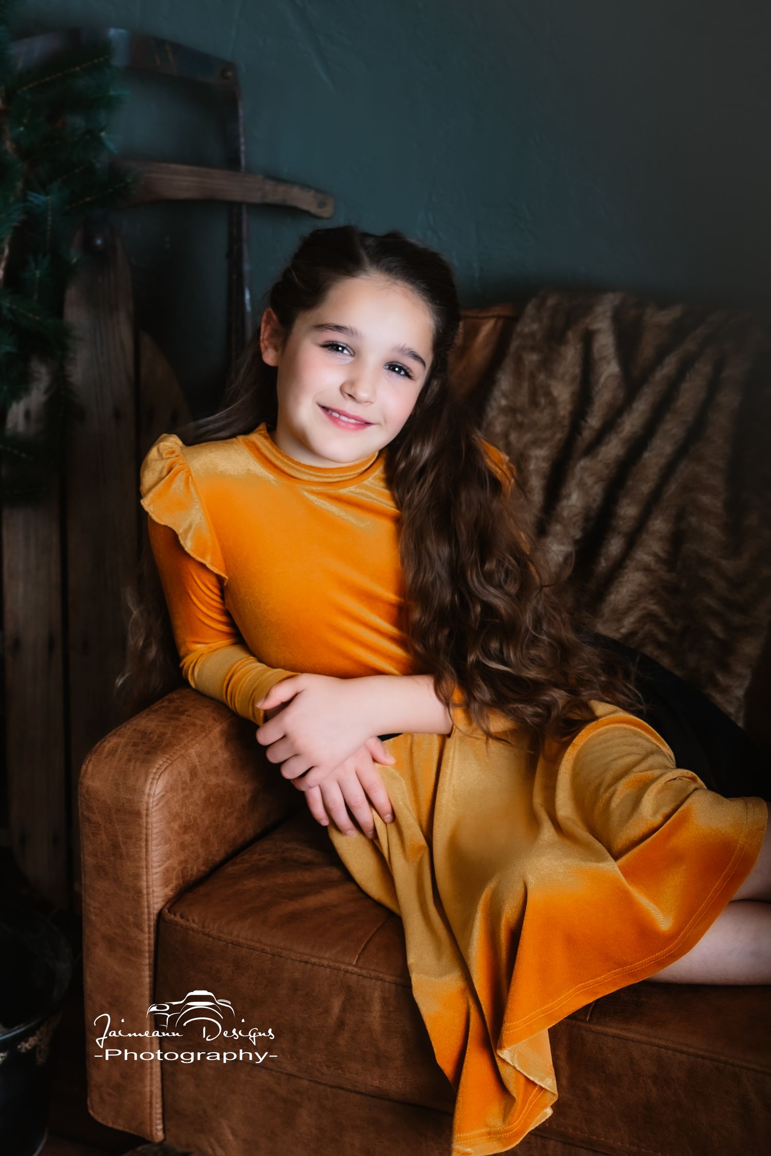 Child in bright orange dress sits gracefully on leather armchair in moody studio setting.