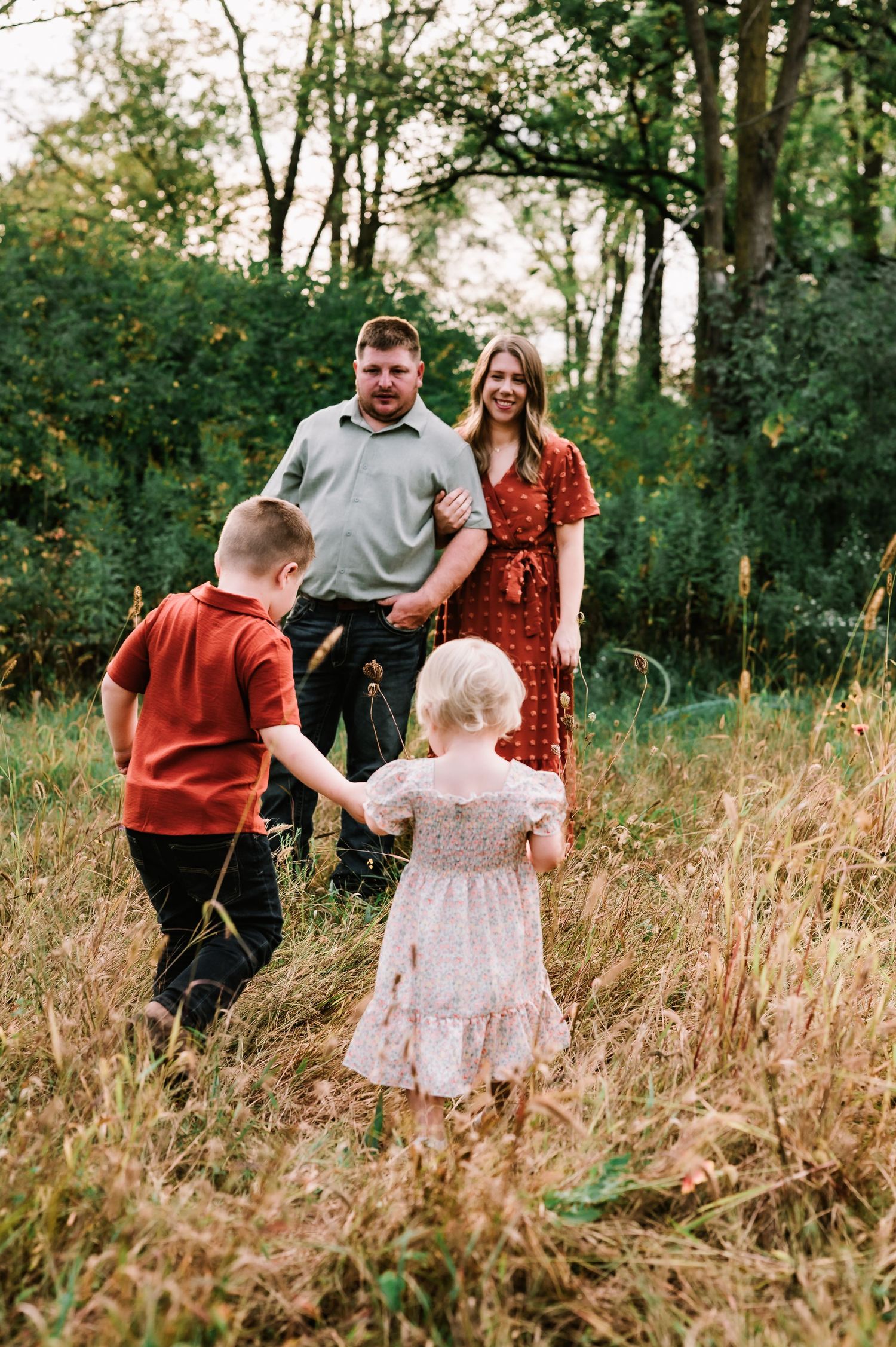 Family of four poses in a grassy meadow surrounded by lush green trees during an outdoor fall photo session.