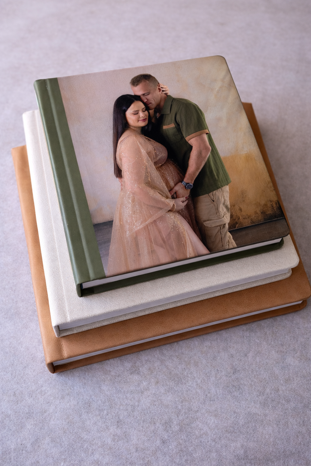 Stacked photo books with a cover image of a pregnant couple embracing, the woman in a pink lace gown and man in green shirt.