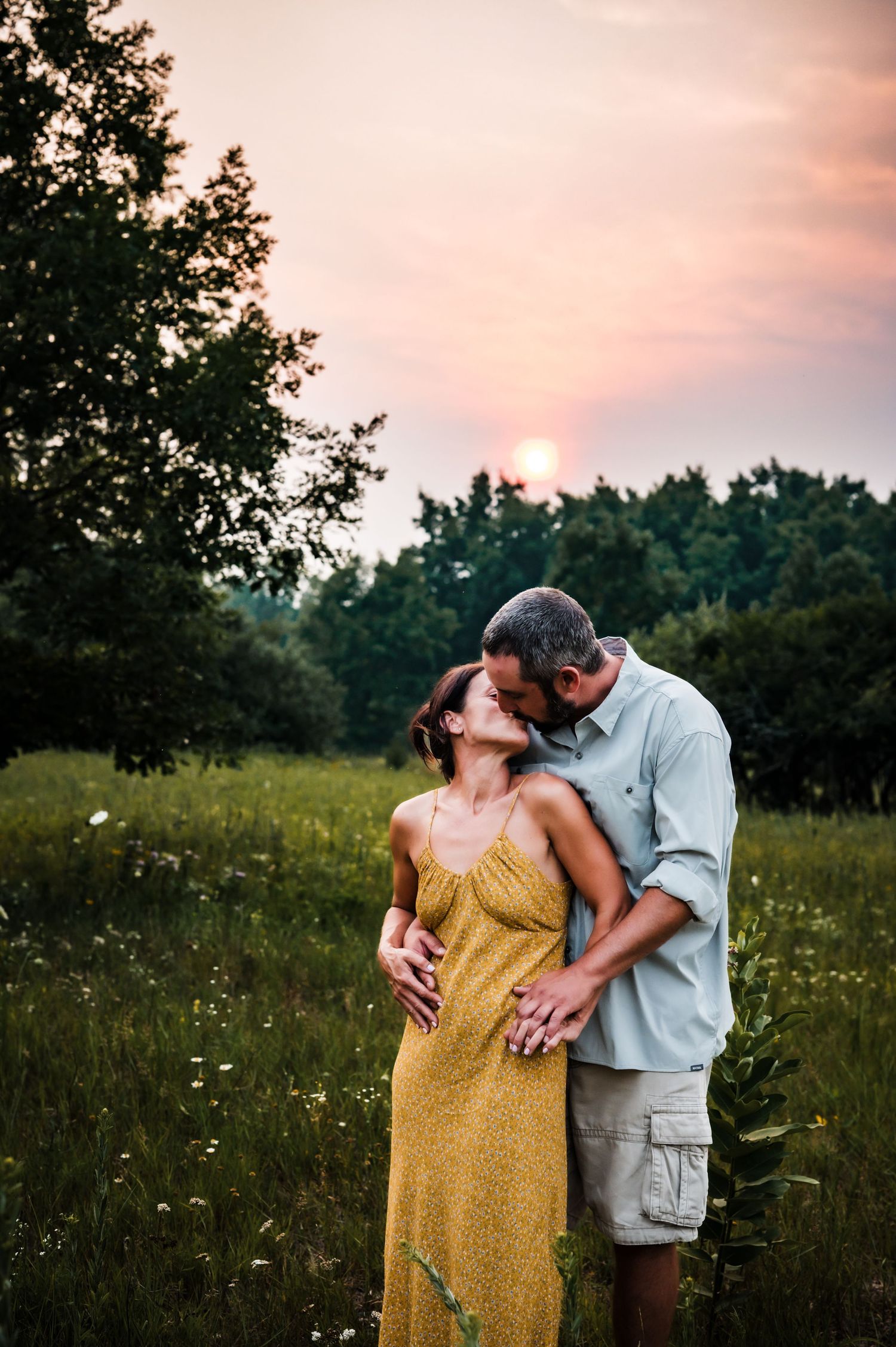 Couple sharing a romantic kiss in a wildflower meadow at golden sunset, woman in yellow dress, surrounded by lush green trees.