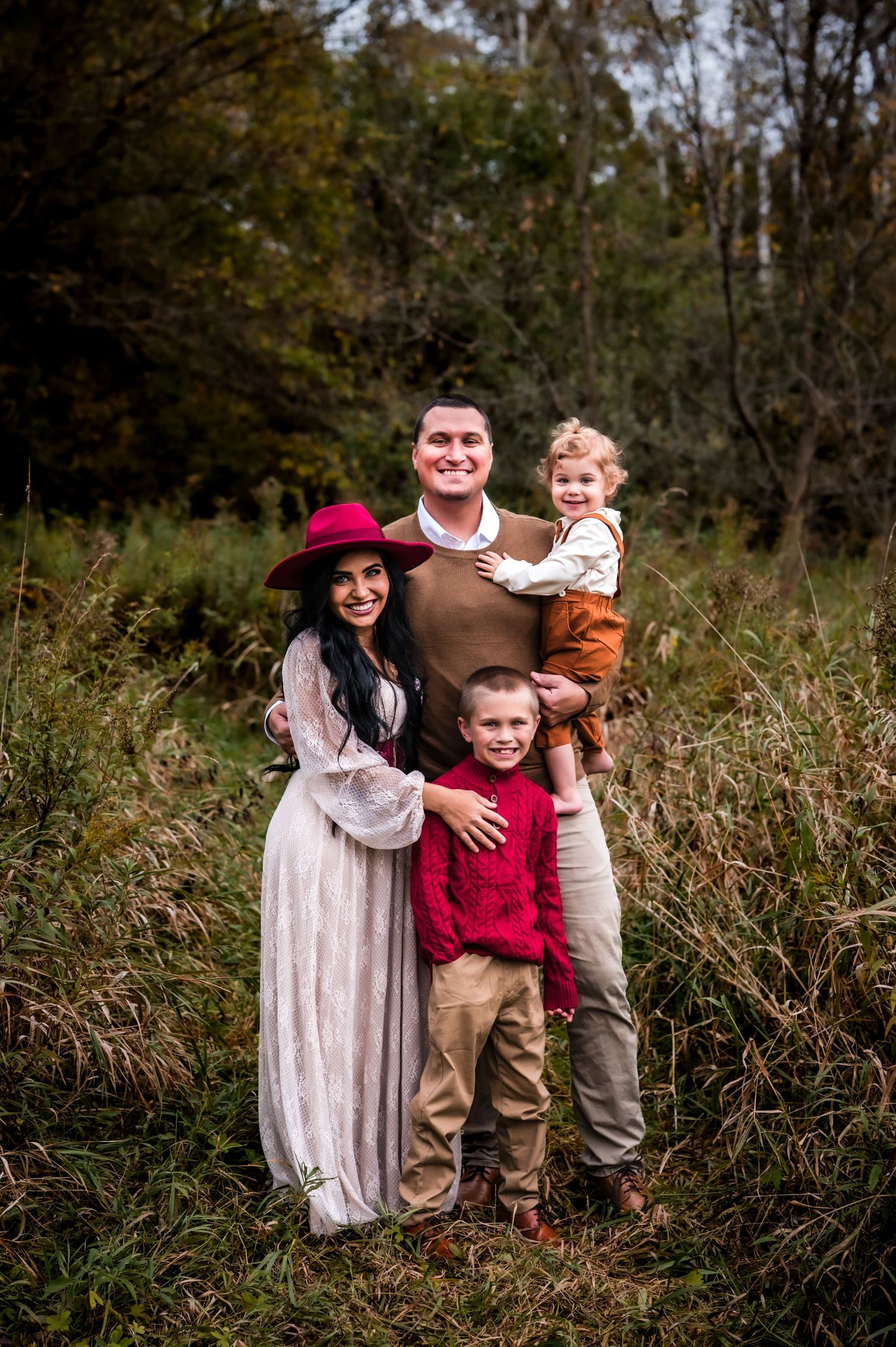 Family of four posing outdoors in autumn field, mom in white lace dress and red hat, dad holding toddler, boy in red sweater.