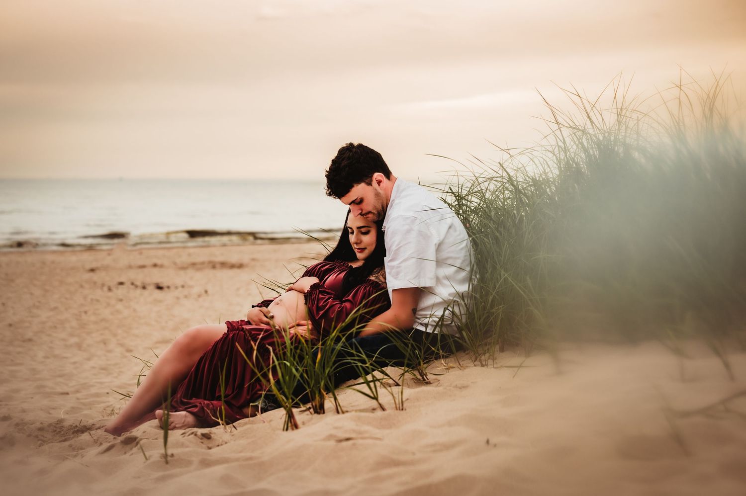 Couple sitting together on sandy beach dunes at sunset, surrounded by tall grass with ocean in background.