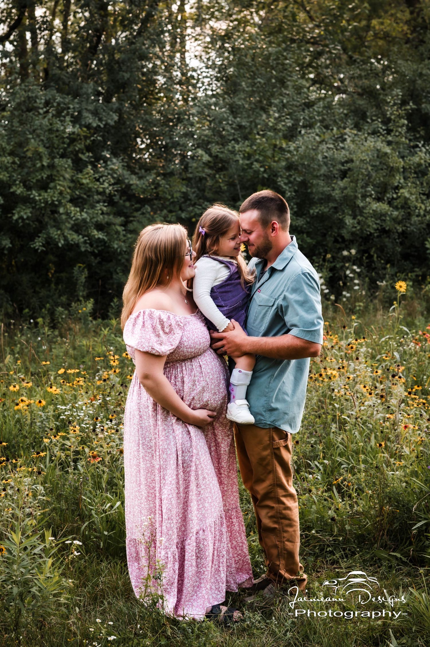 Pregnant woman in pink floral gown with partner holding child in sunny wildflower meadow, maternity photography.