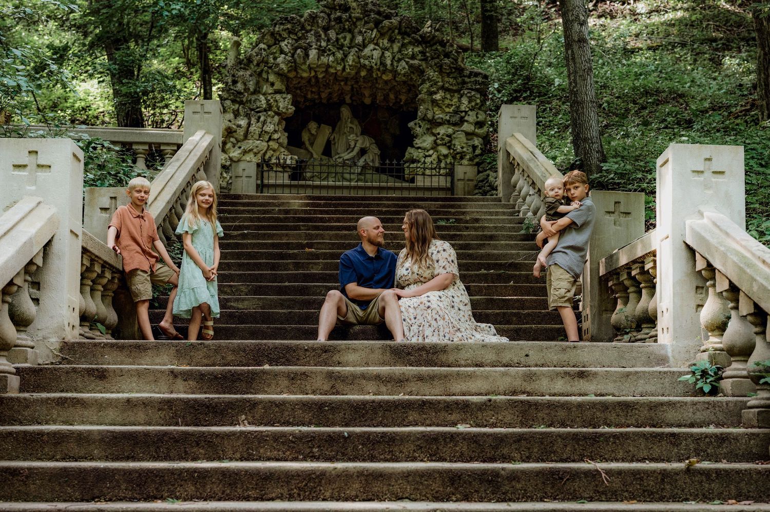 Family posing on ornate stone steps in a lush garden, with parents seated centrally and children standing beside them.