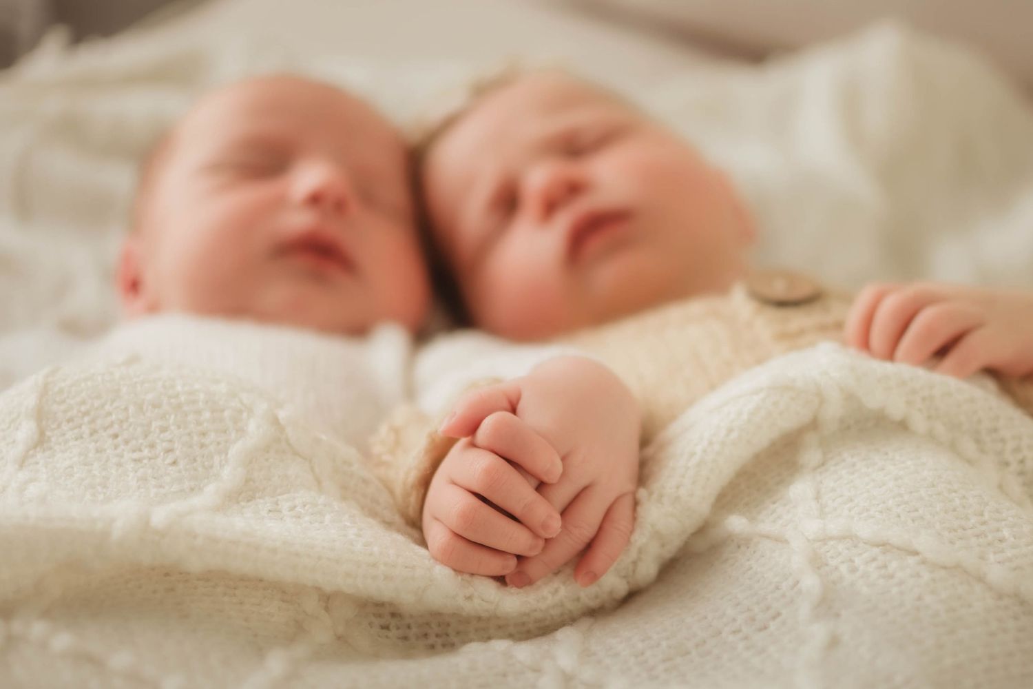 Two sleeping newborn twins lie side by side wrapped in a soft cream knit blanket, with their tiny hands clasped together in the foreground.