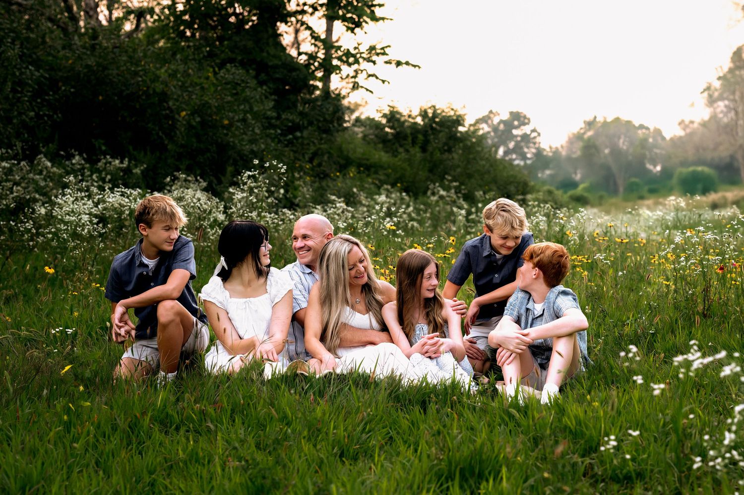 A group of people relax together in a lush meadow filled with white wildflowers during a summer evening private outdoor studio, WI