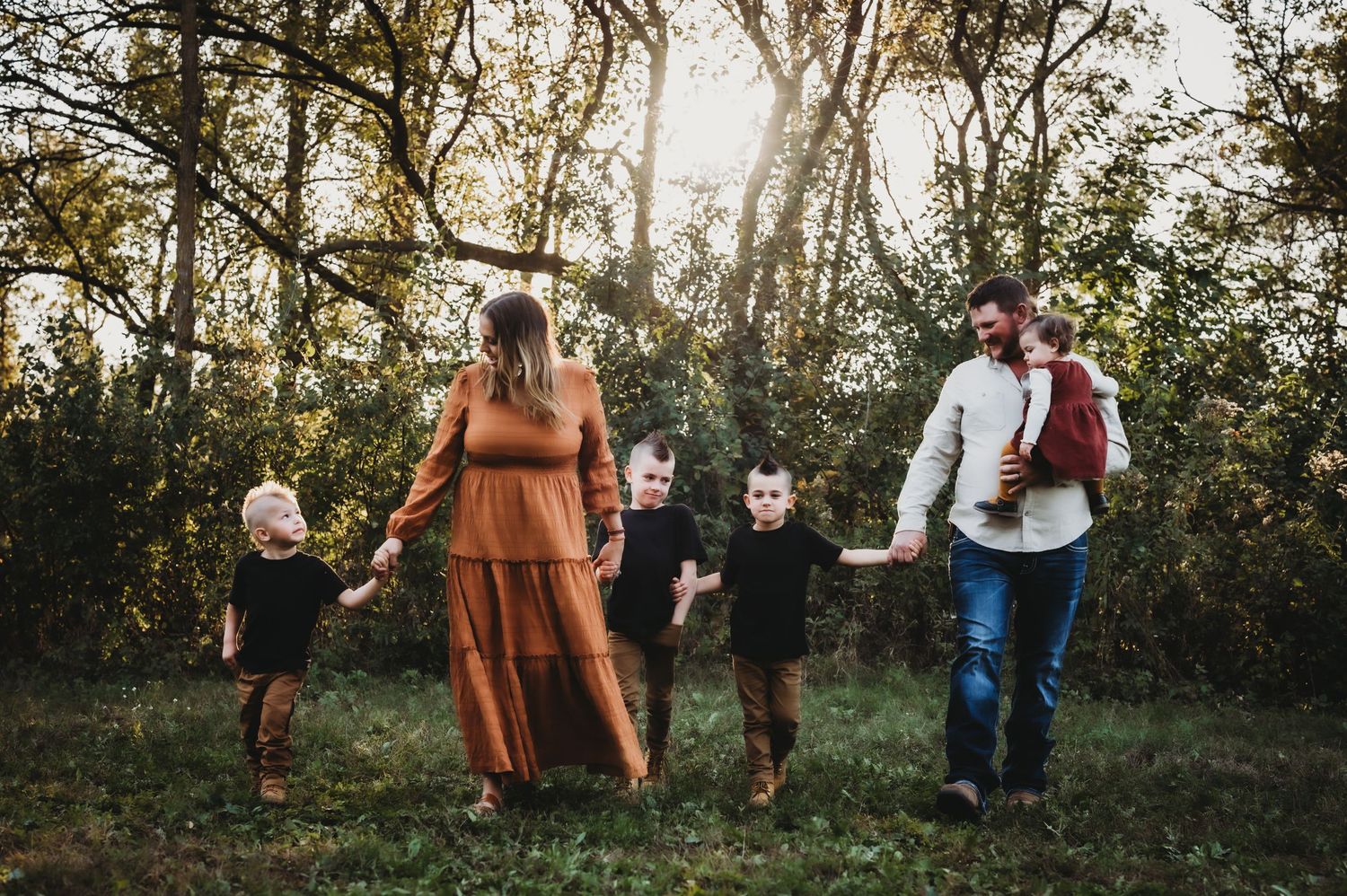 A family walks together through a wooded area at sunset, dressed in warm autumn colors and casual attire.
