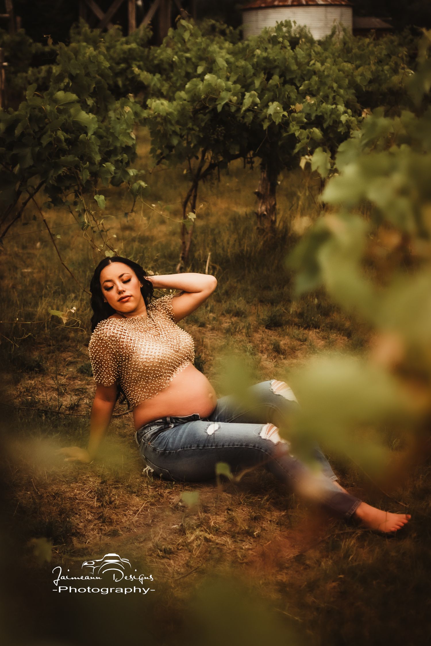 Relaxed maternity pose among grapevines wearing sparkly top and jeans.