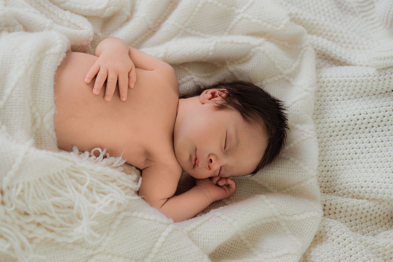 Peaceful newborn baby sleeping on a soft white knit blanket with dark hair and tiny hands tucked under cheek.