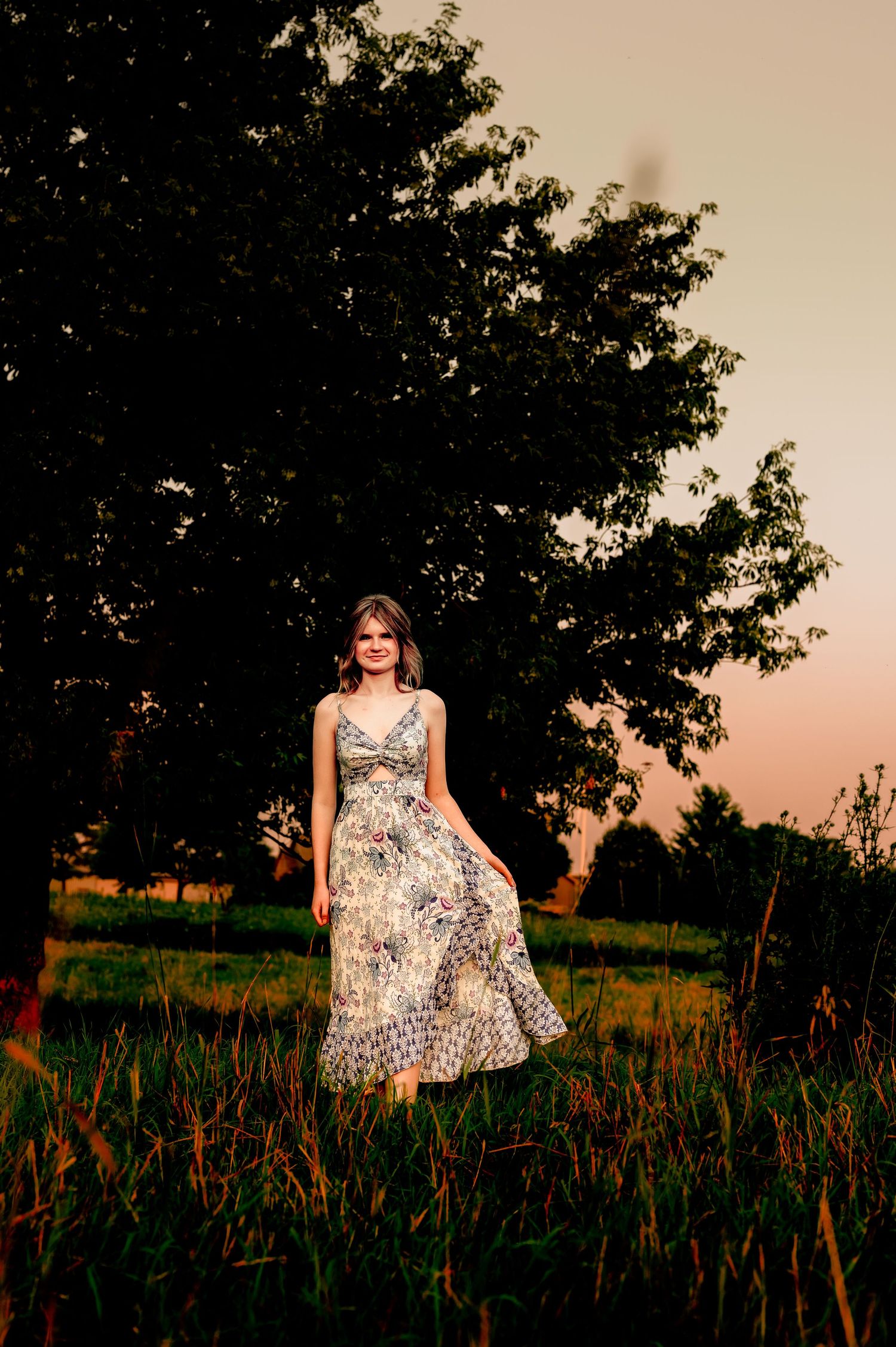 A figure in a floral dress twirls in a grassy field at sunset with trees silhouetted in the background in Eden, WI