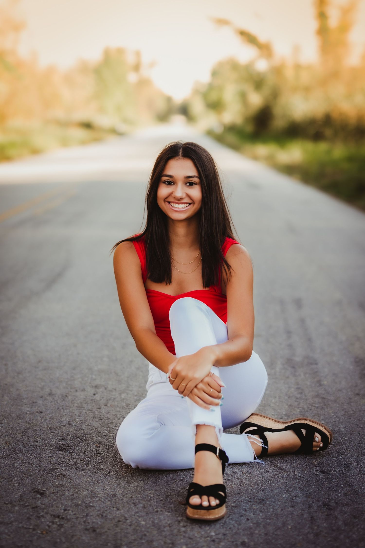 Person in red top and white pants sits on a rural road at sunset wearing sandals in Campbellsport, WI