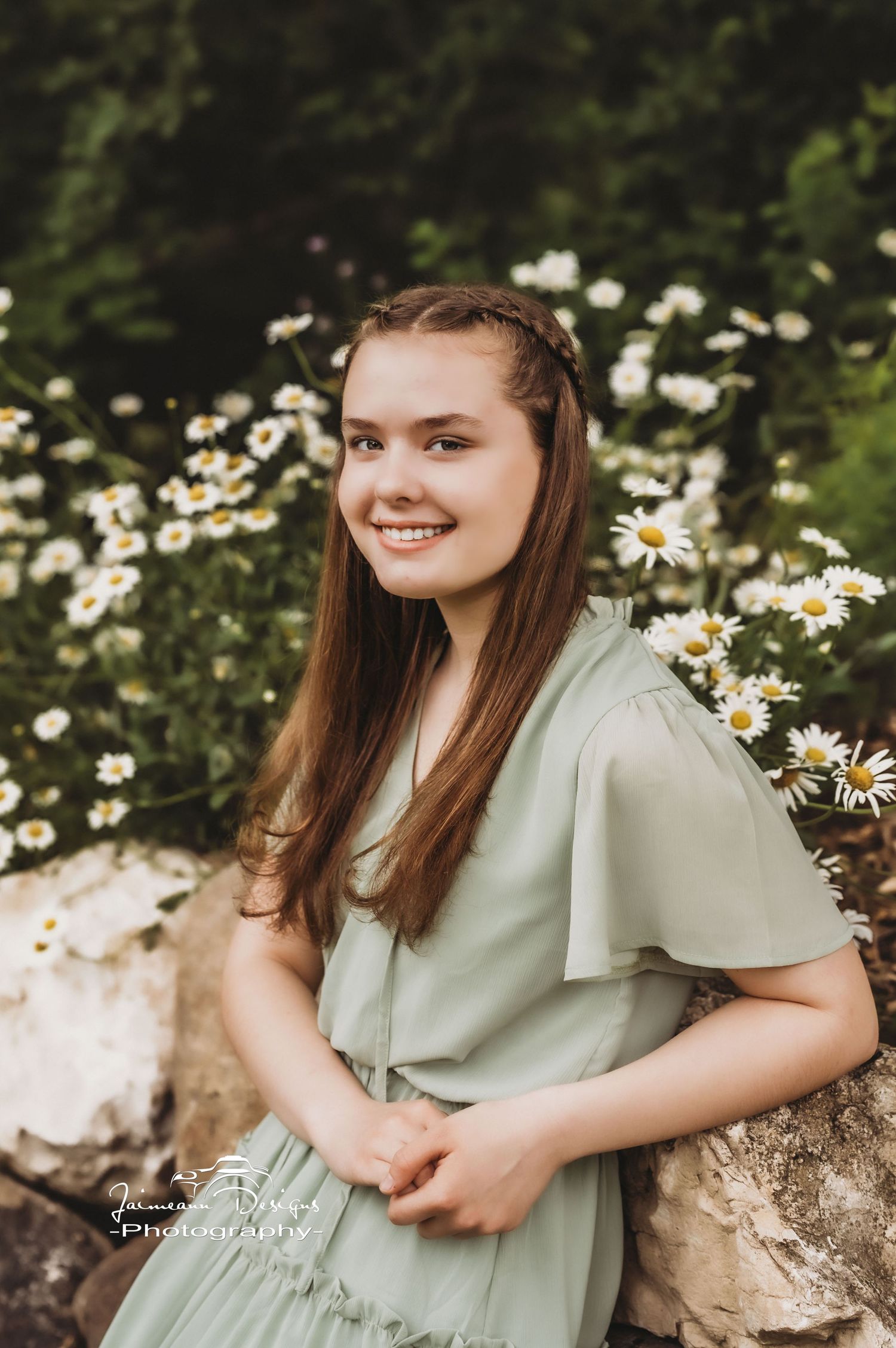 A person in a sage green dress poses by white daisies in a natural outdoor setting.