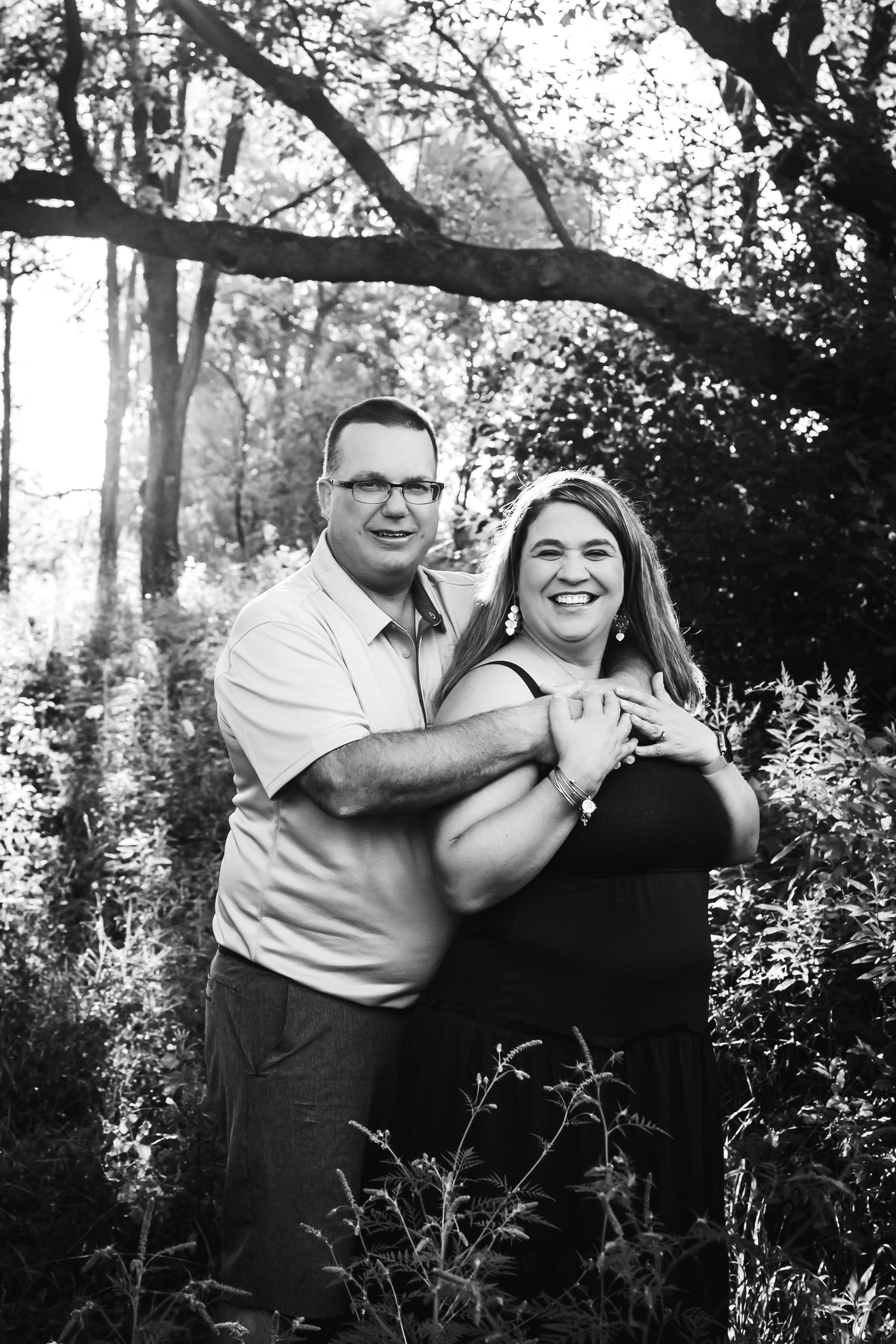 A romantic couple embraces while laughing together in a wooded outdoor setting with natural lighting.