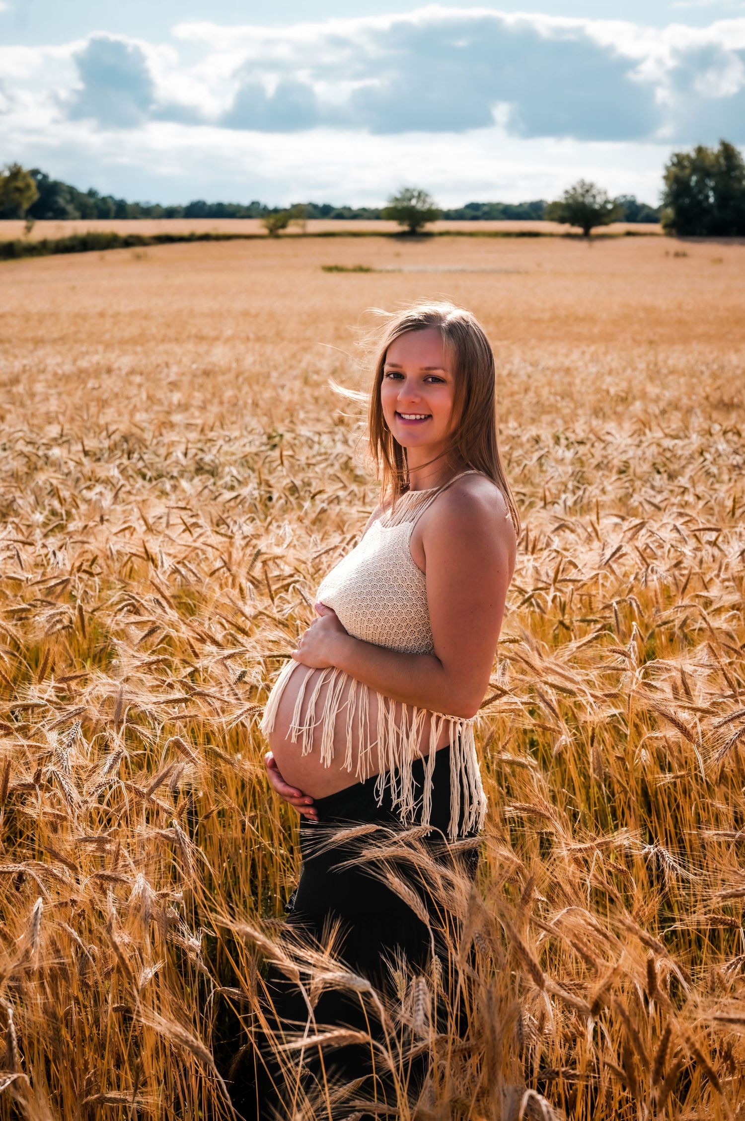 Smiling pregnant woman in a white fringed crop top standing in a golden wheat field on a sunny day.