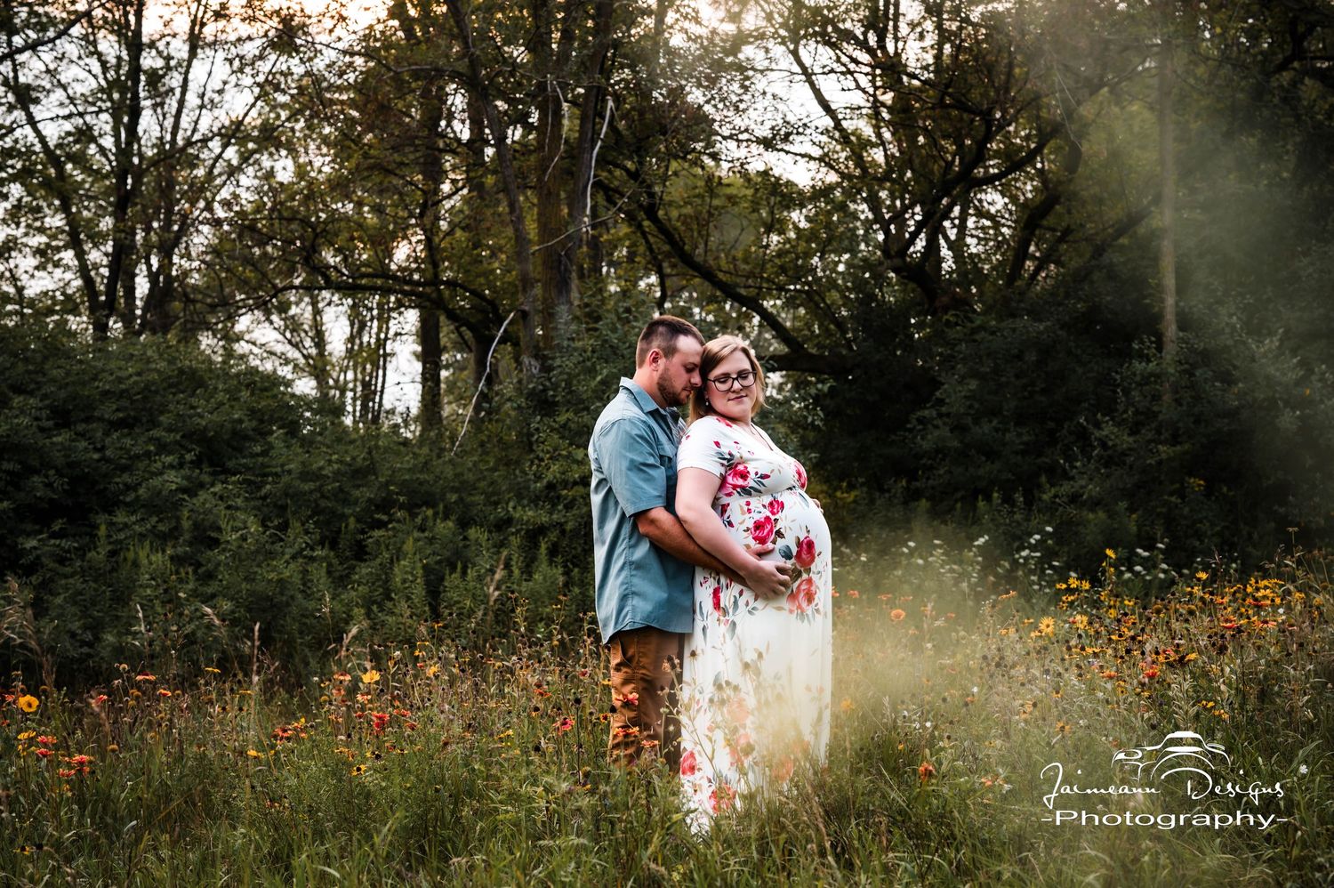 Couple embracing in a sunlit wildflower meadow during a maternity photo session, surrounded by lush green trees.
