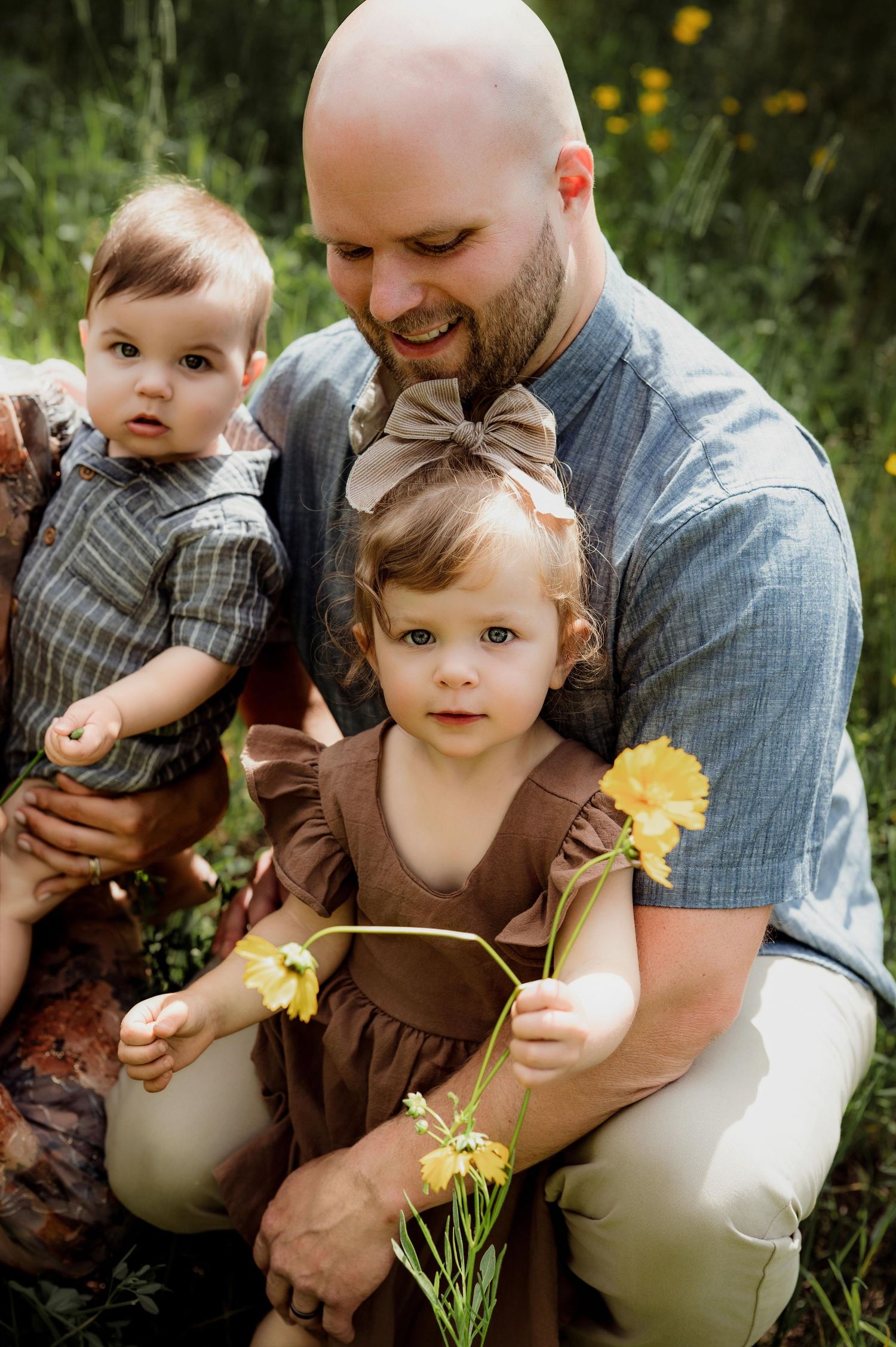 Bald father kneeling outdoors holding baby boy while toddler girl in brown dress holds yellow wildflowers.