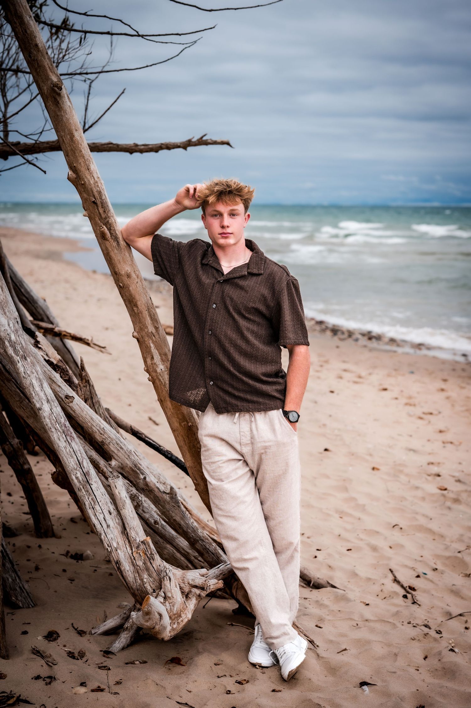 Young man in brown shirt and beige pants leaning against driftwood on a sandy beach with ocean waves behind him.
