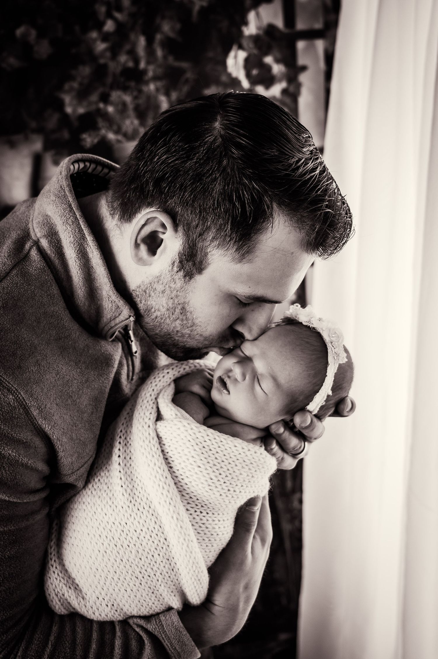 Black and white portrait of tender moment between parent and swaddled newborn in knit blanket.