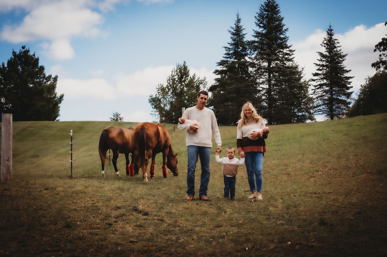 Family walks together through a field with horses grazing in the background under blue skies.