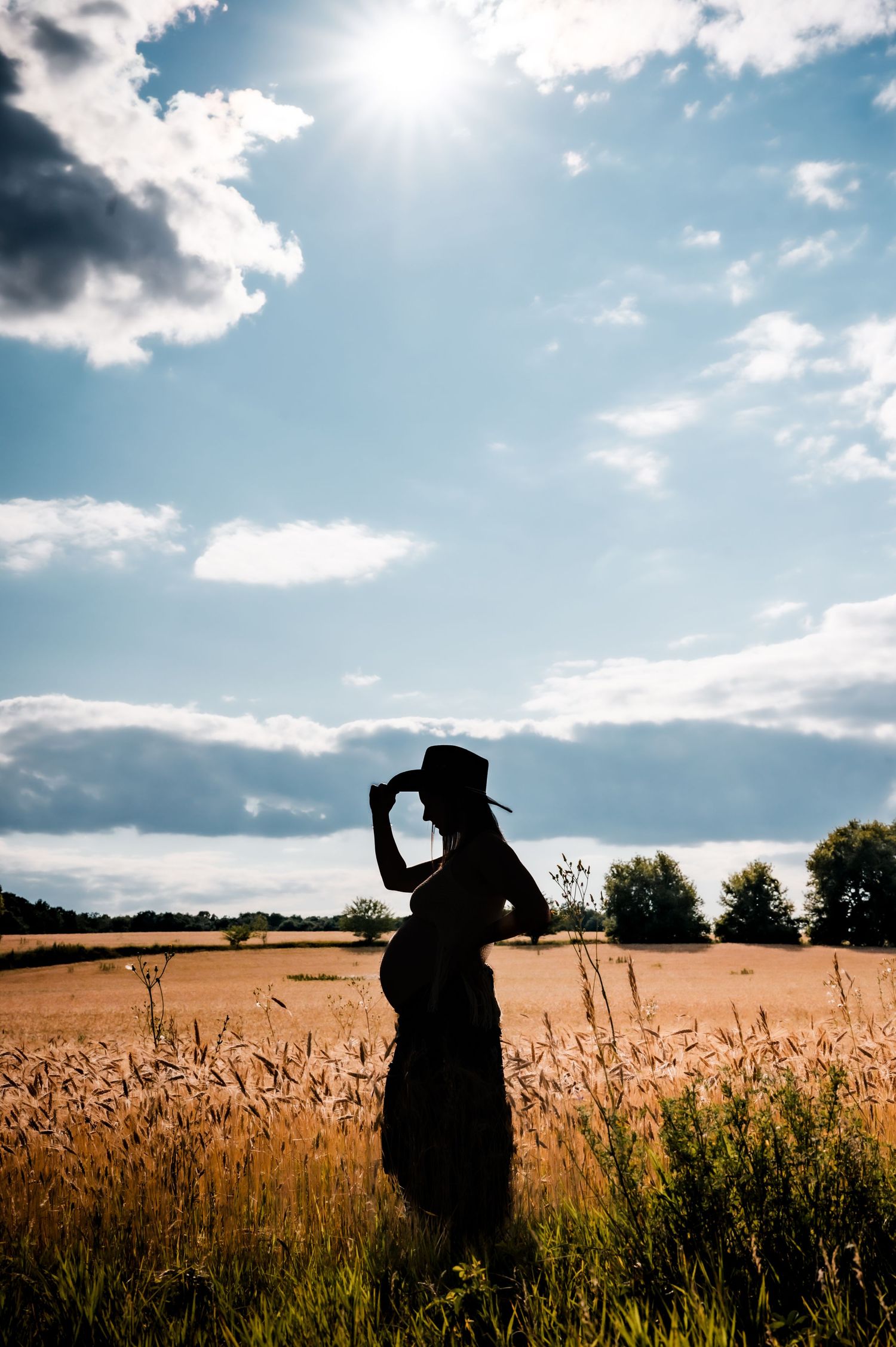 Silhouette of a pregnant woman in a cowboy hat standing in a golden wheat field under a dramatic sunny sky.