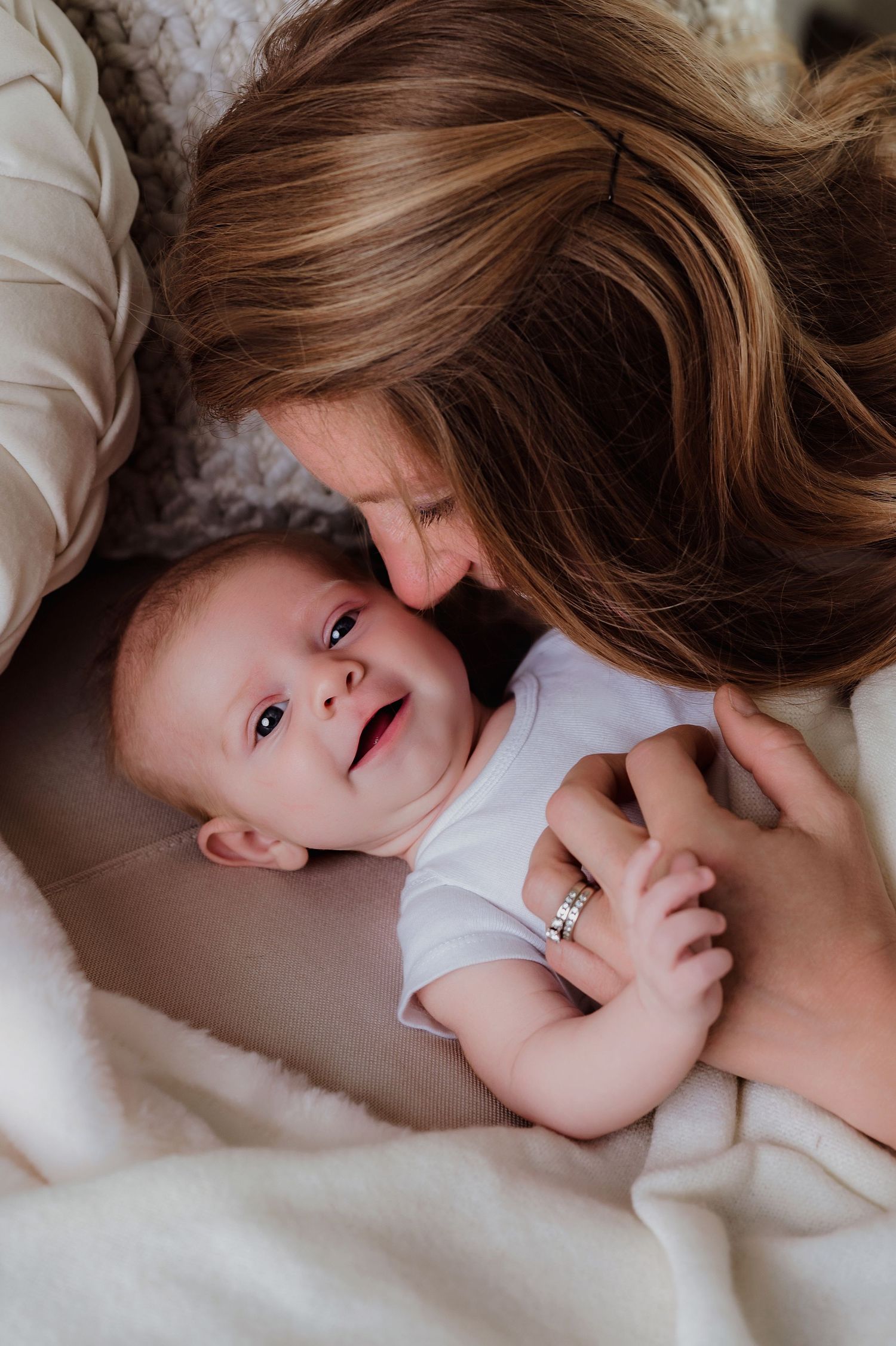 A mother tenderly nuzzles her smiling baby lying on a white blanket, wearing a white onesie.