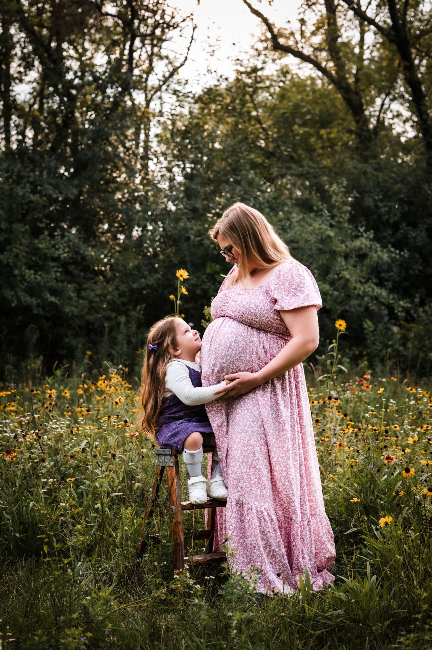 Pregnant woman in pink floral dress with young daughter in wildflower field during maternity photo session.
