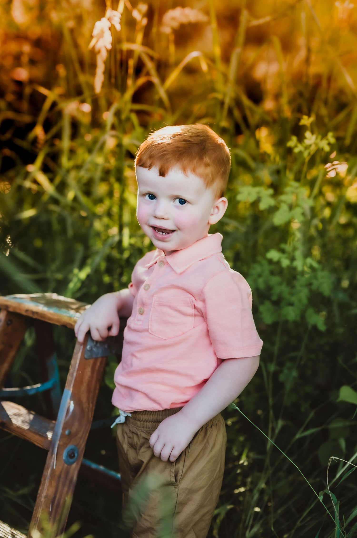 Young child in pink polo shirt smiling while standing by wooden railing in natural setting.