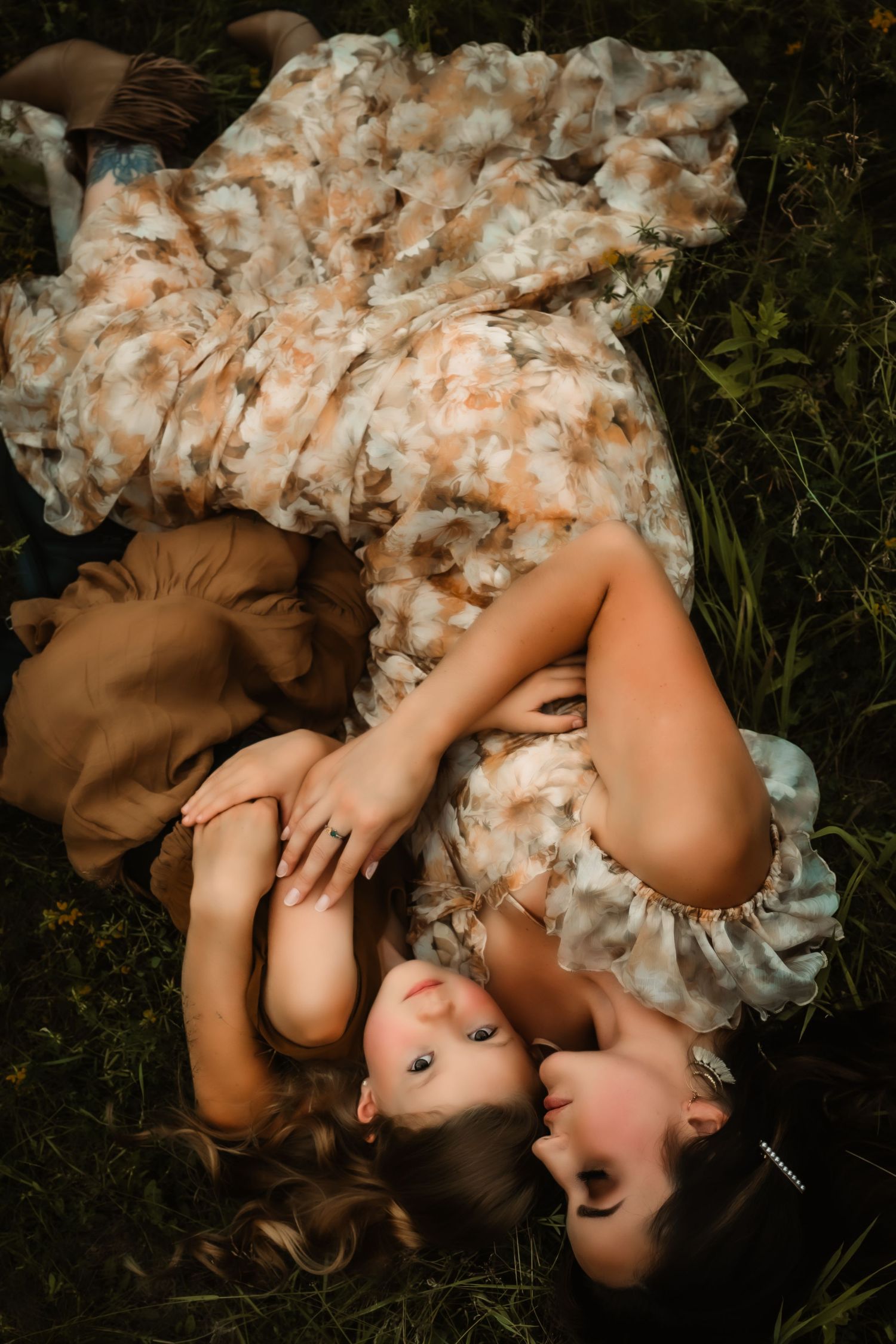 Two people lying in grass wearing floral dresses during sunset photoshoot.