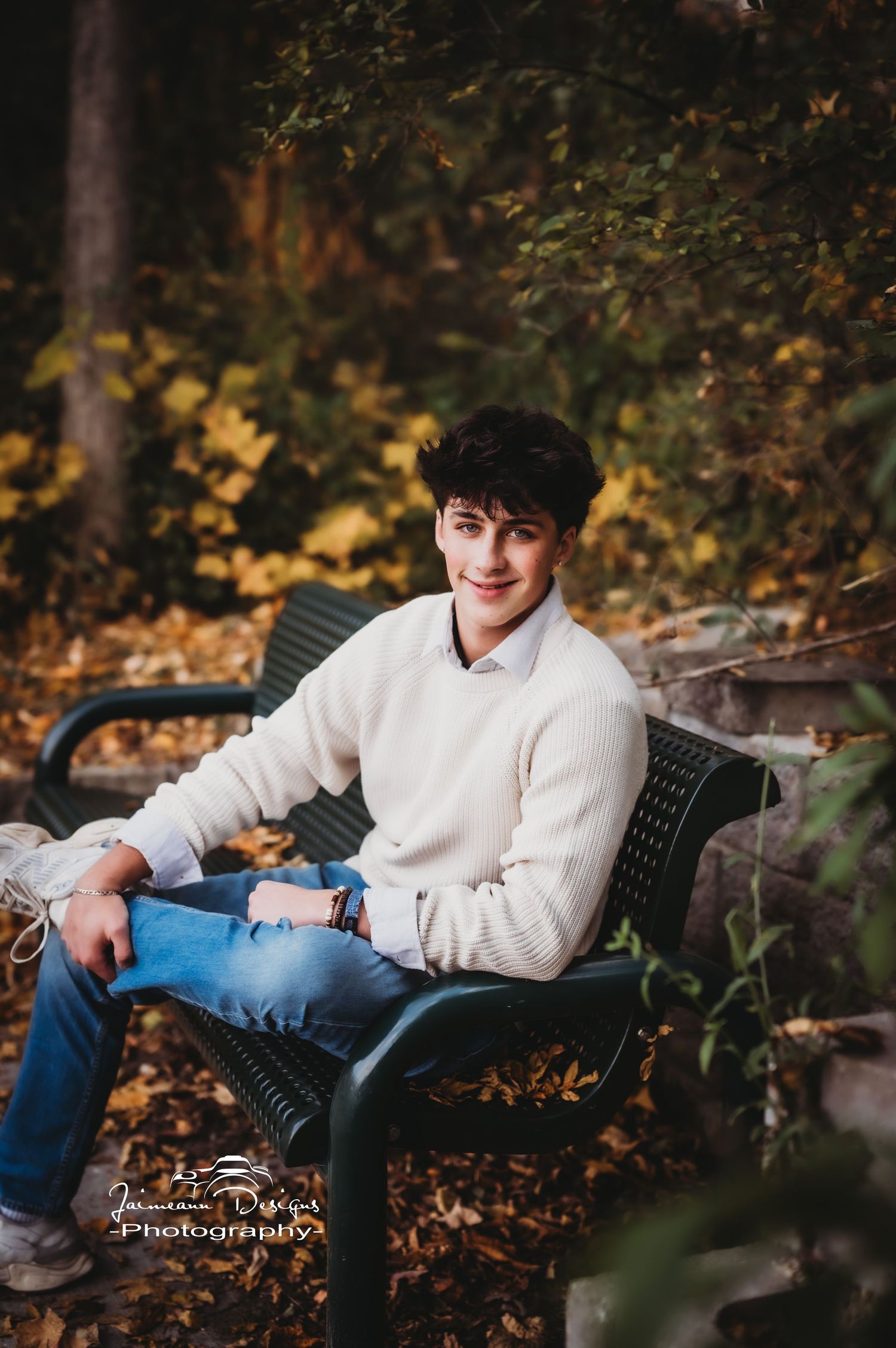 Portrait on park bench wearing cream sweater with fall foliage backdrop.