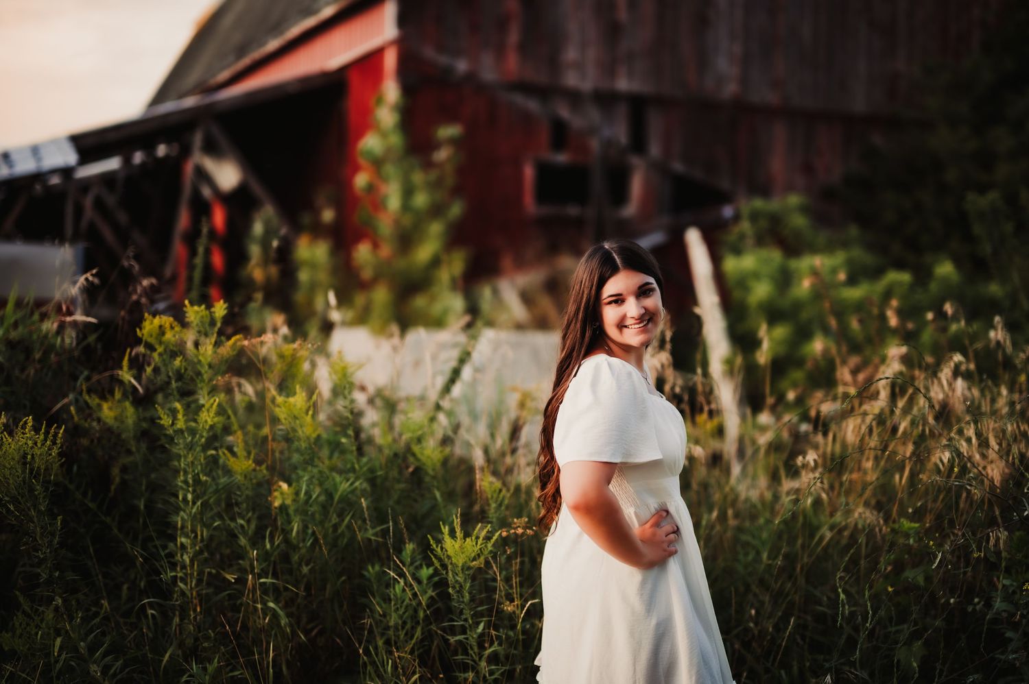 White dress flows in evening light with rustic red barn backdrop.