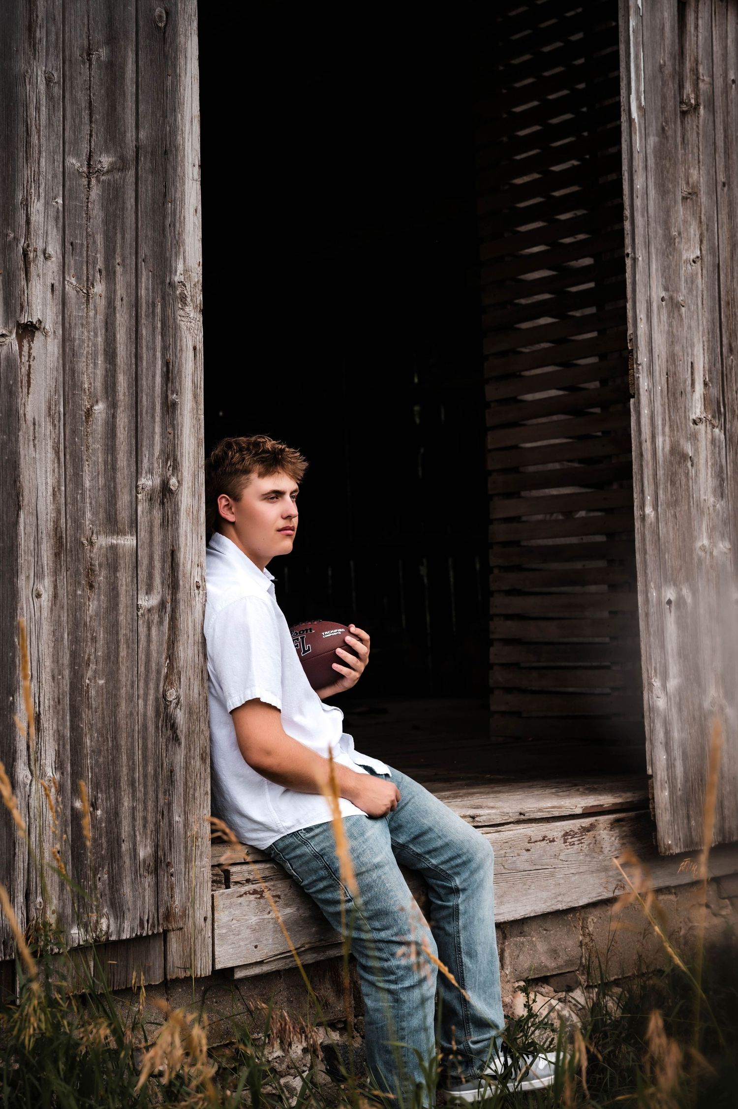 Teen boy in white shirt and jeans holds football while sitting in rustic barn doorway.
