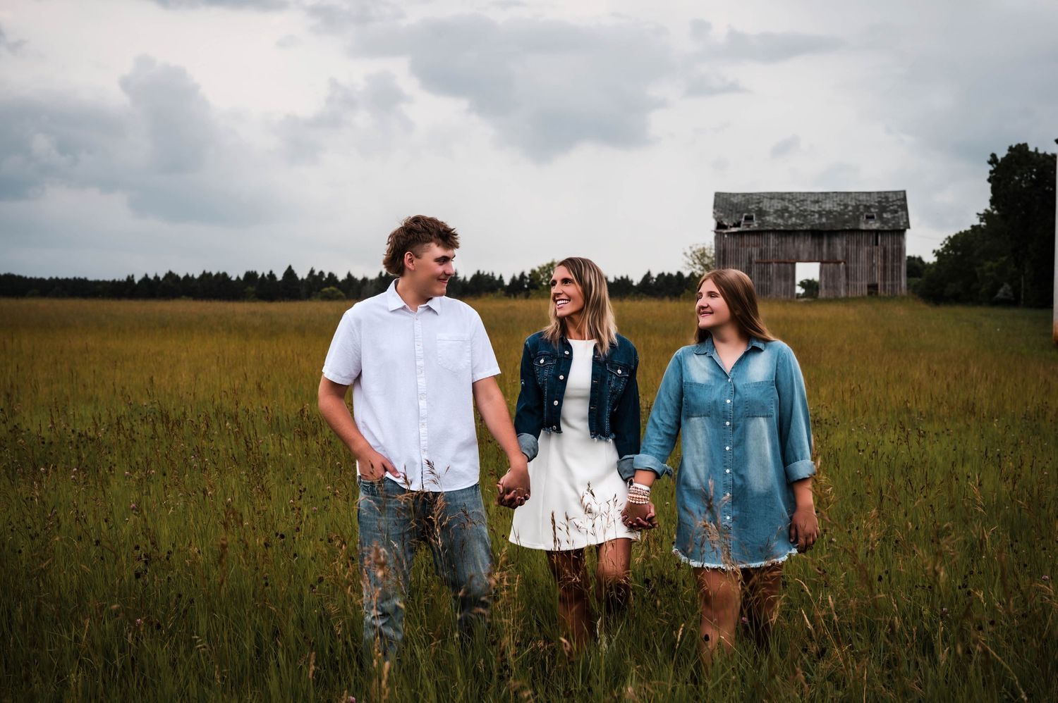 Three people holding hands smiling in a green field with a rustic barn in the background on a cloudy day.