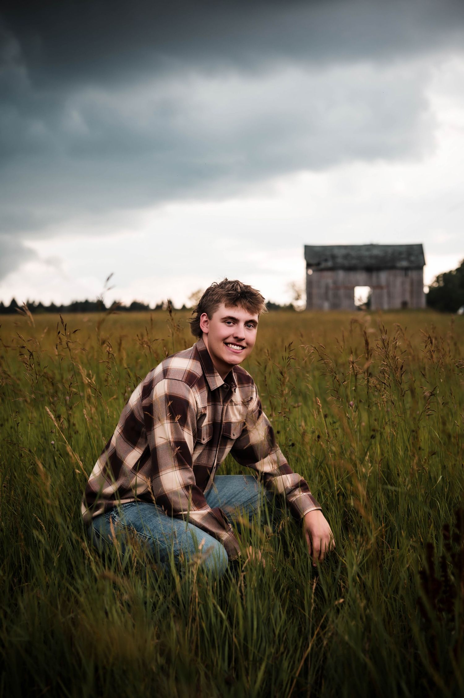 Young man in plaid shirt crouching in tall green field with rustic barn and stormy sky backdrop.