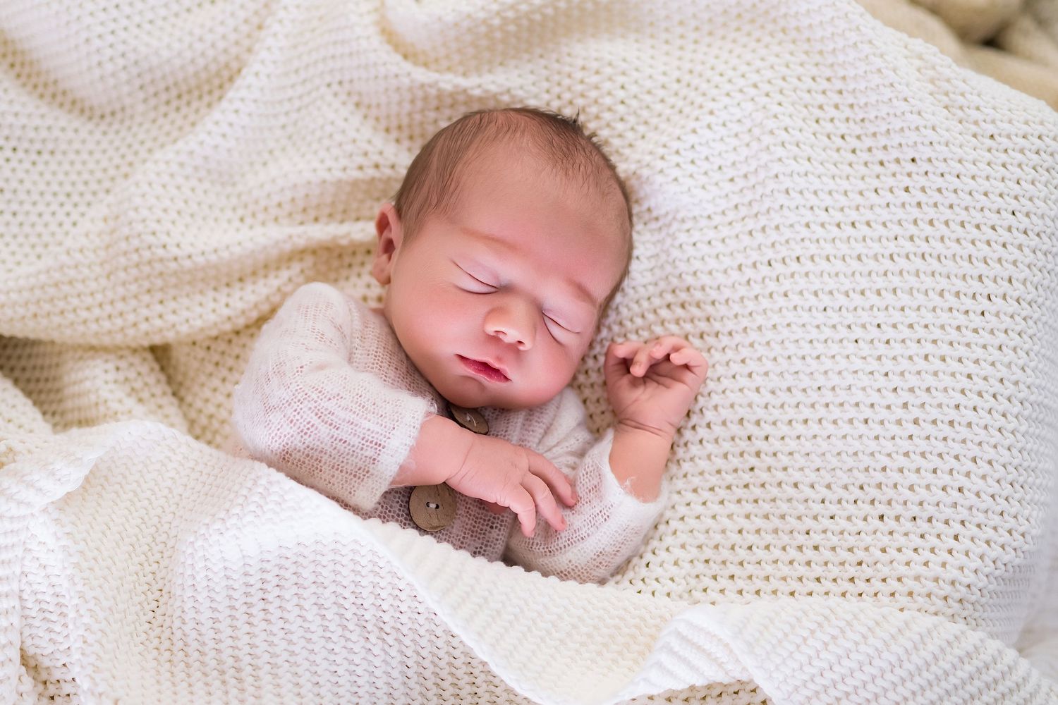 Peaceful newborn baby sleeping on soft cream knit blanket wearing a cozy white outfit.