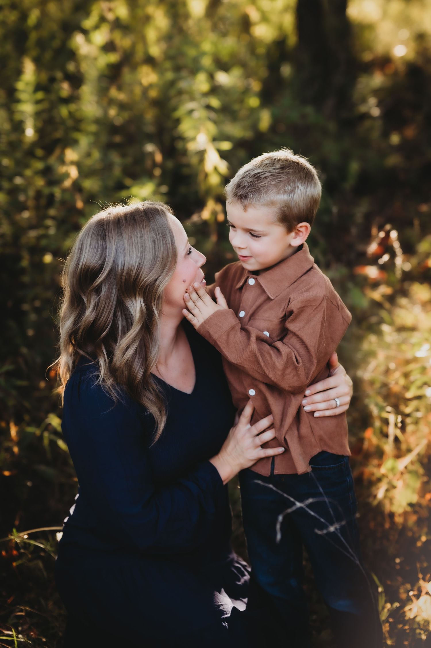 A mother and young boy share a tender moment outdoors, surrounded by lush green foliage in warm autumn light.