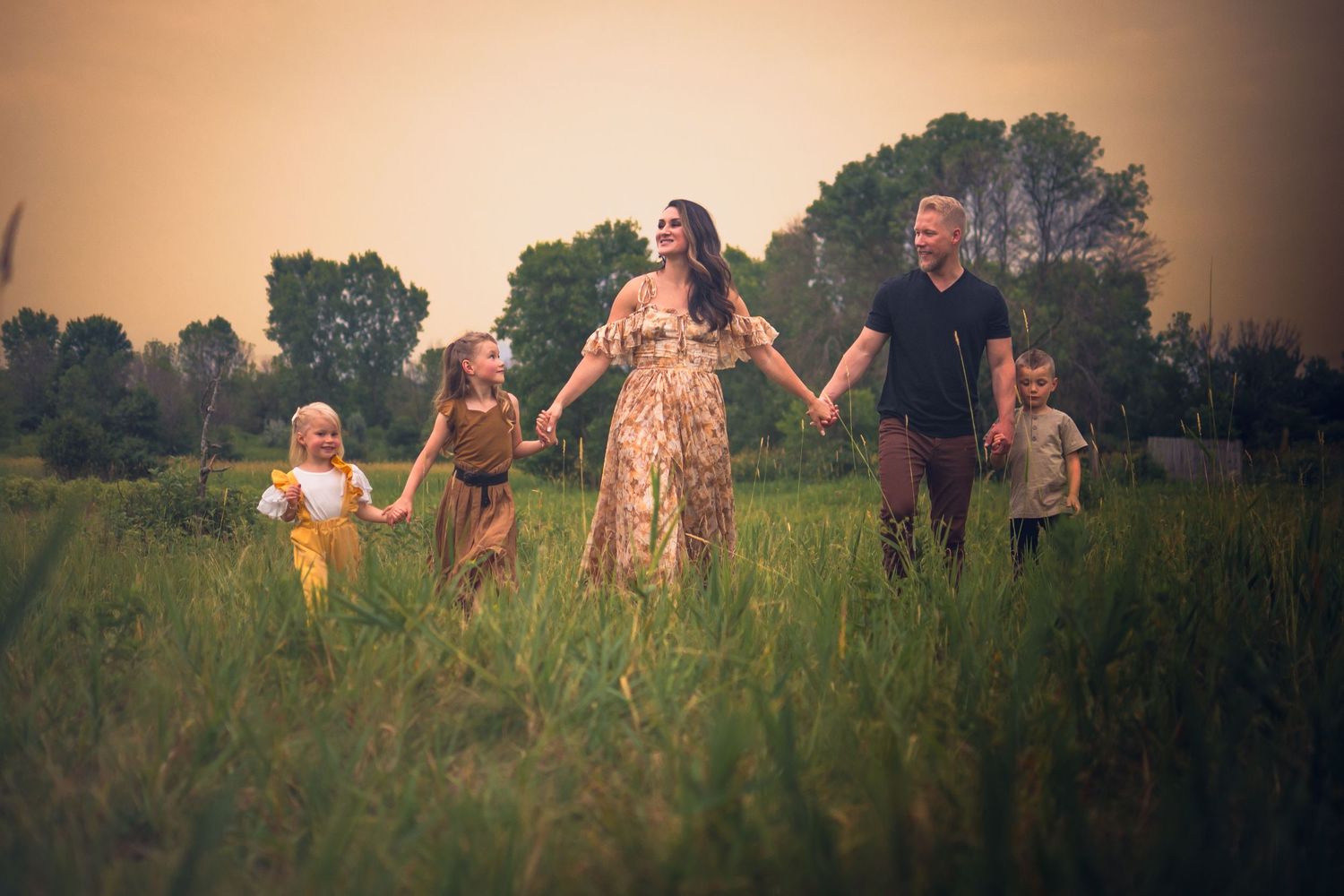 Family holding hands while walking through tall grass meadow at sunset.