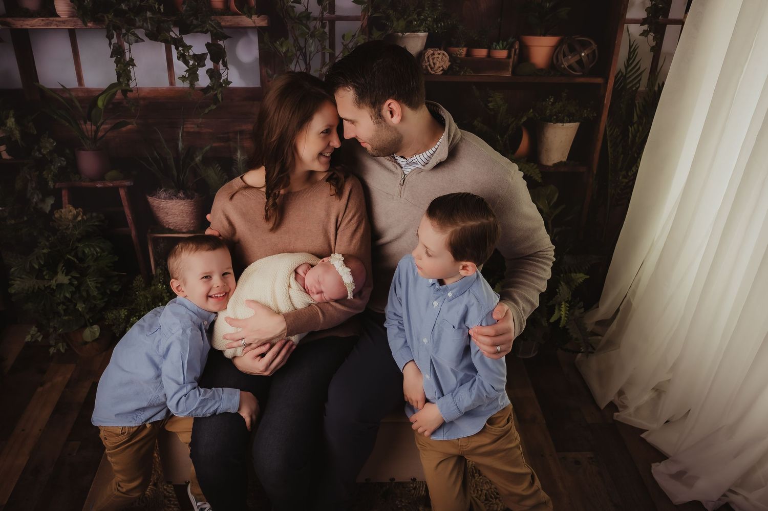 Warm family portrait featuring parents with young children and newborn surrounded by indoor plants.