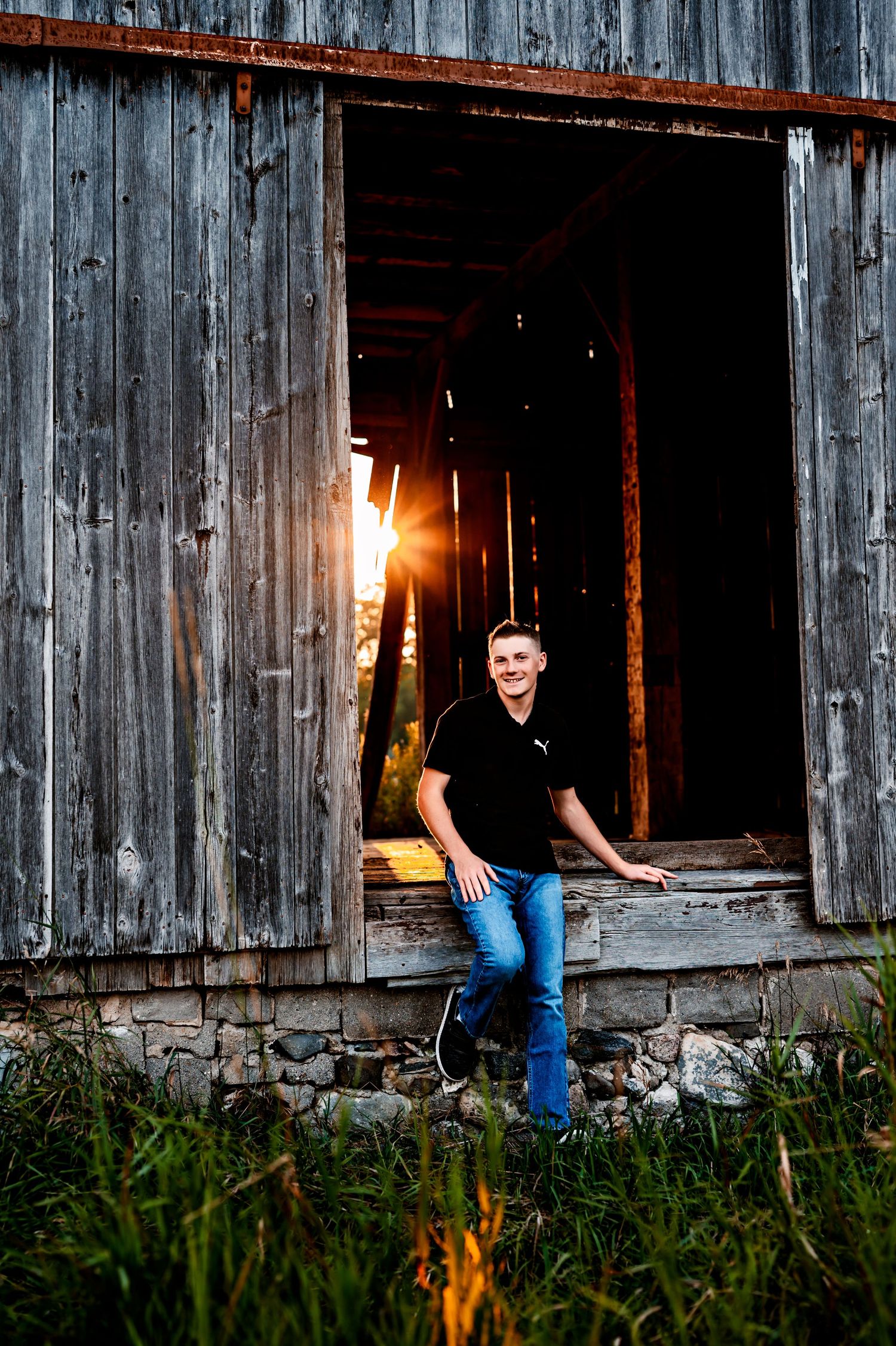Person sits casually on wooden barn foundation at sunset with weathered gray boards and tall grass in foreground in Allenton, WI