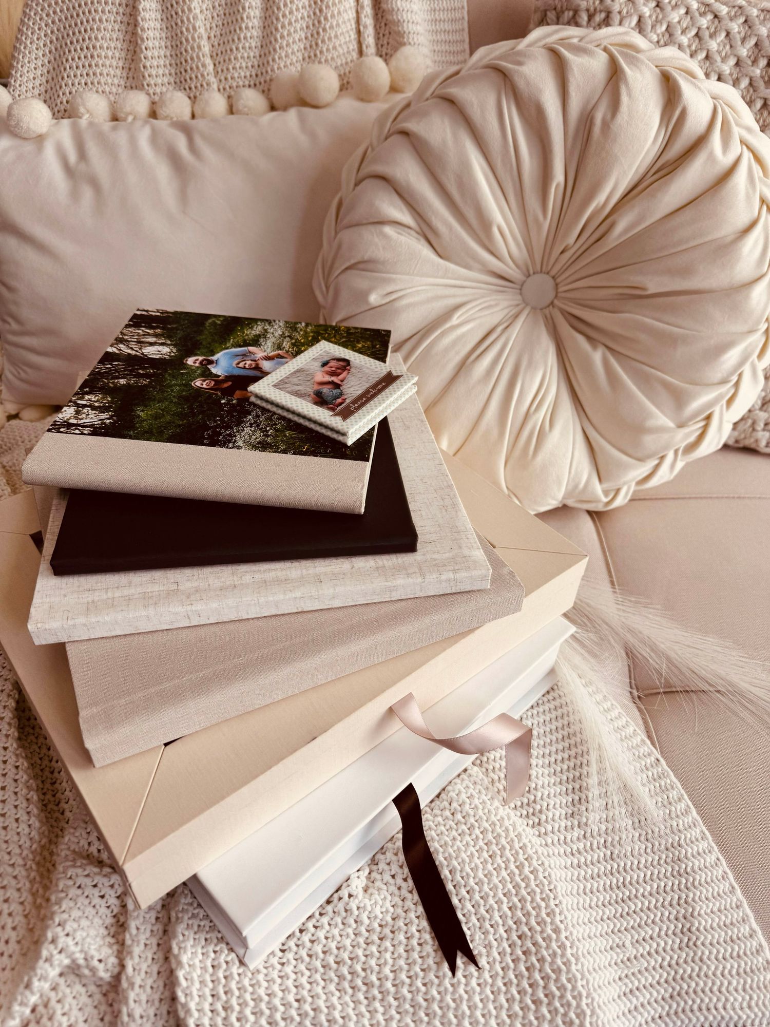 Cream velvet round pillow beside stacked photo books on a cozy neutral sofa with knit throw blanket.