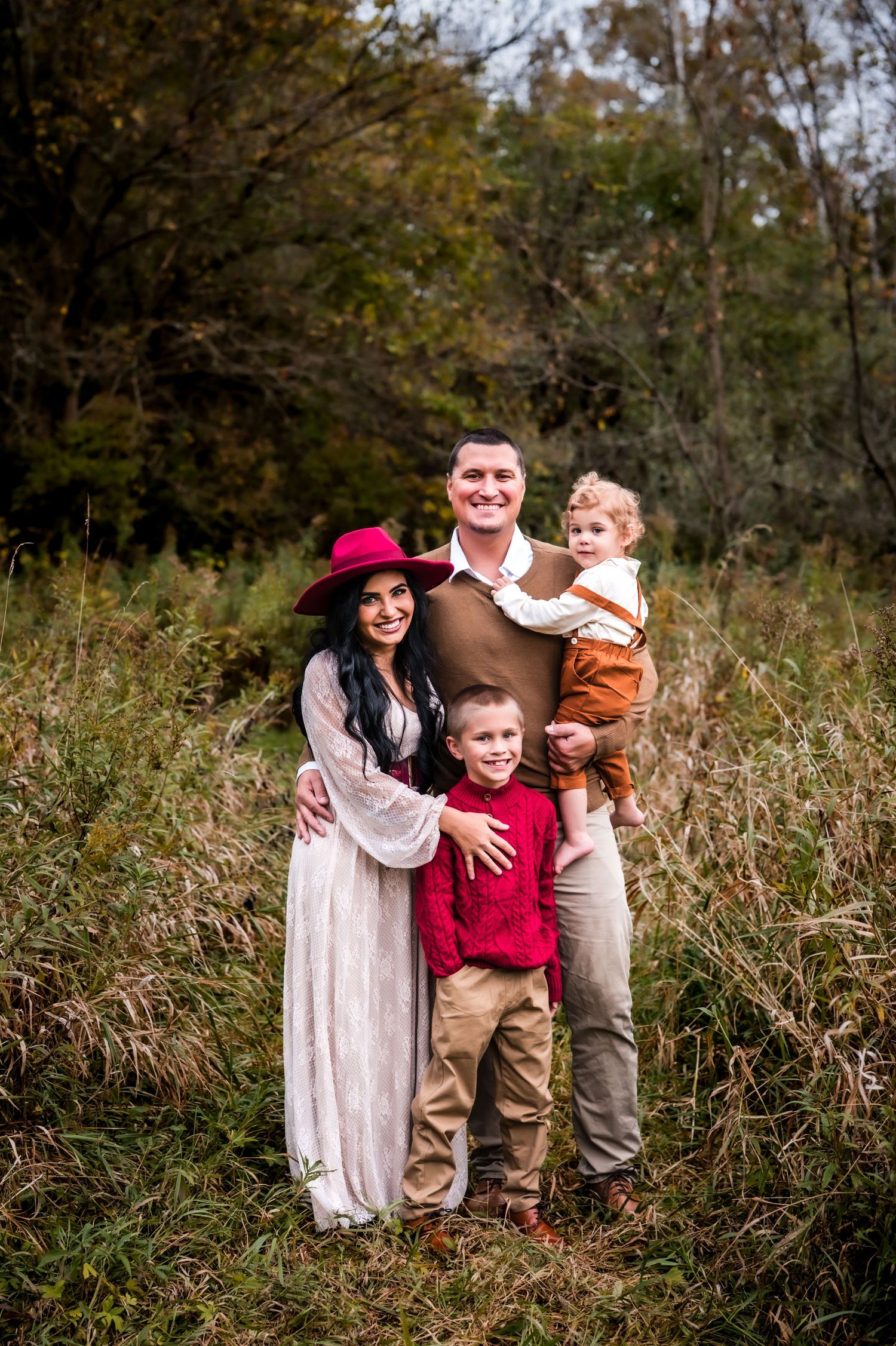 Family of four posing outdoors in autumn field, mother in white dress and red hat, father holding toddler, young boy in red sweater.